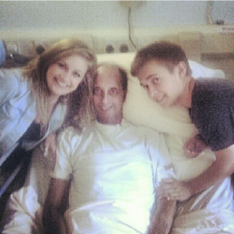 A man lying in bed with two children, a girl on his left and a boy on his right, all smiling for a selfie.