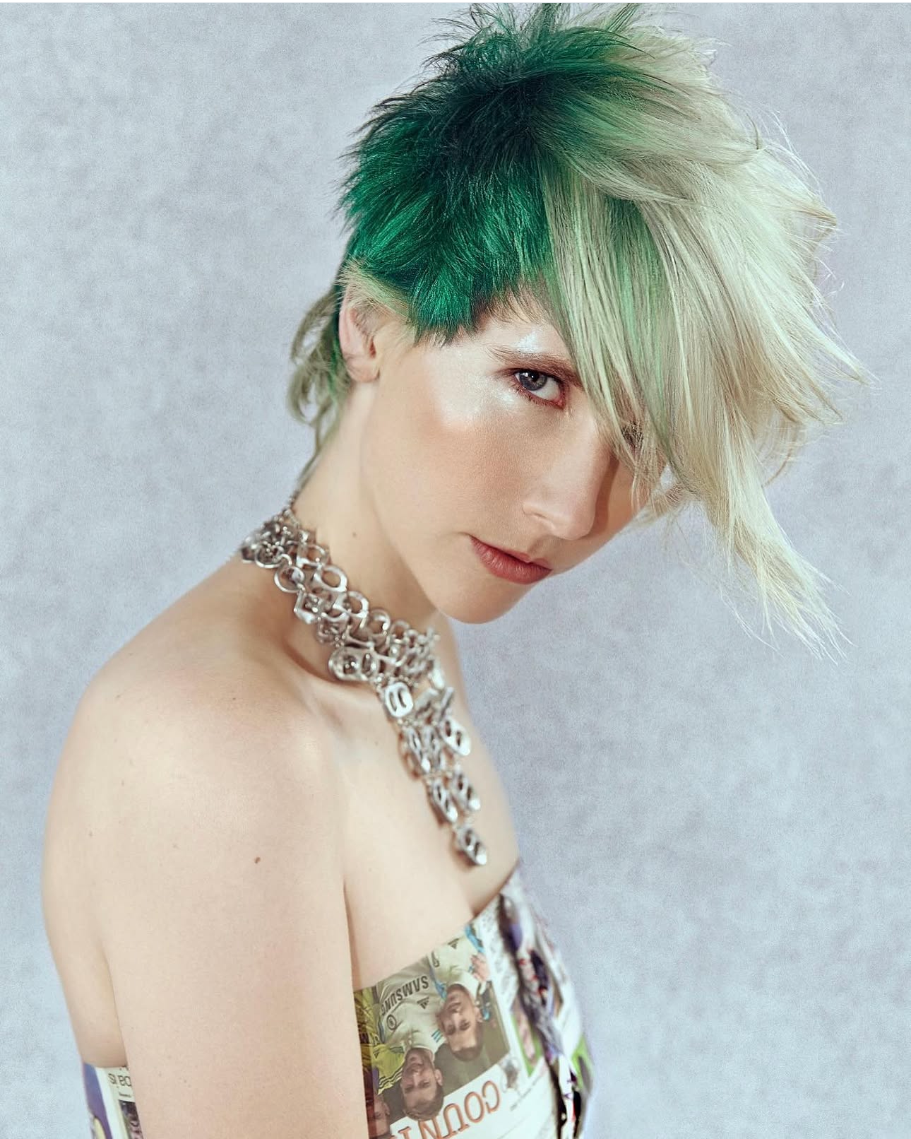A person with short, spiky hair dyed green and platinum blonde, wearing a chunky silver chain necklace and a top made from printed currency notes, looking at the camera with a serious expression against a gray background.