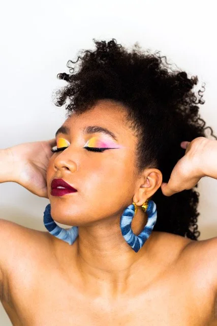 Woman with curly hair and colorful makeup, wearing blue earrings, eyes closed, hands behind head.