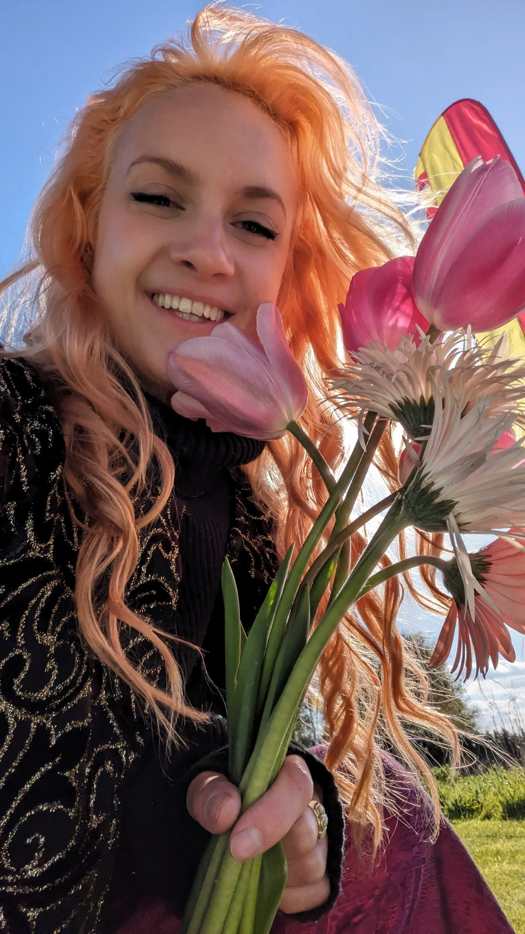 A woman with long curly peach-colored hair holding a bouquet of pink and white flowers, smiling outdoors with a blue sky background.