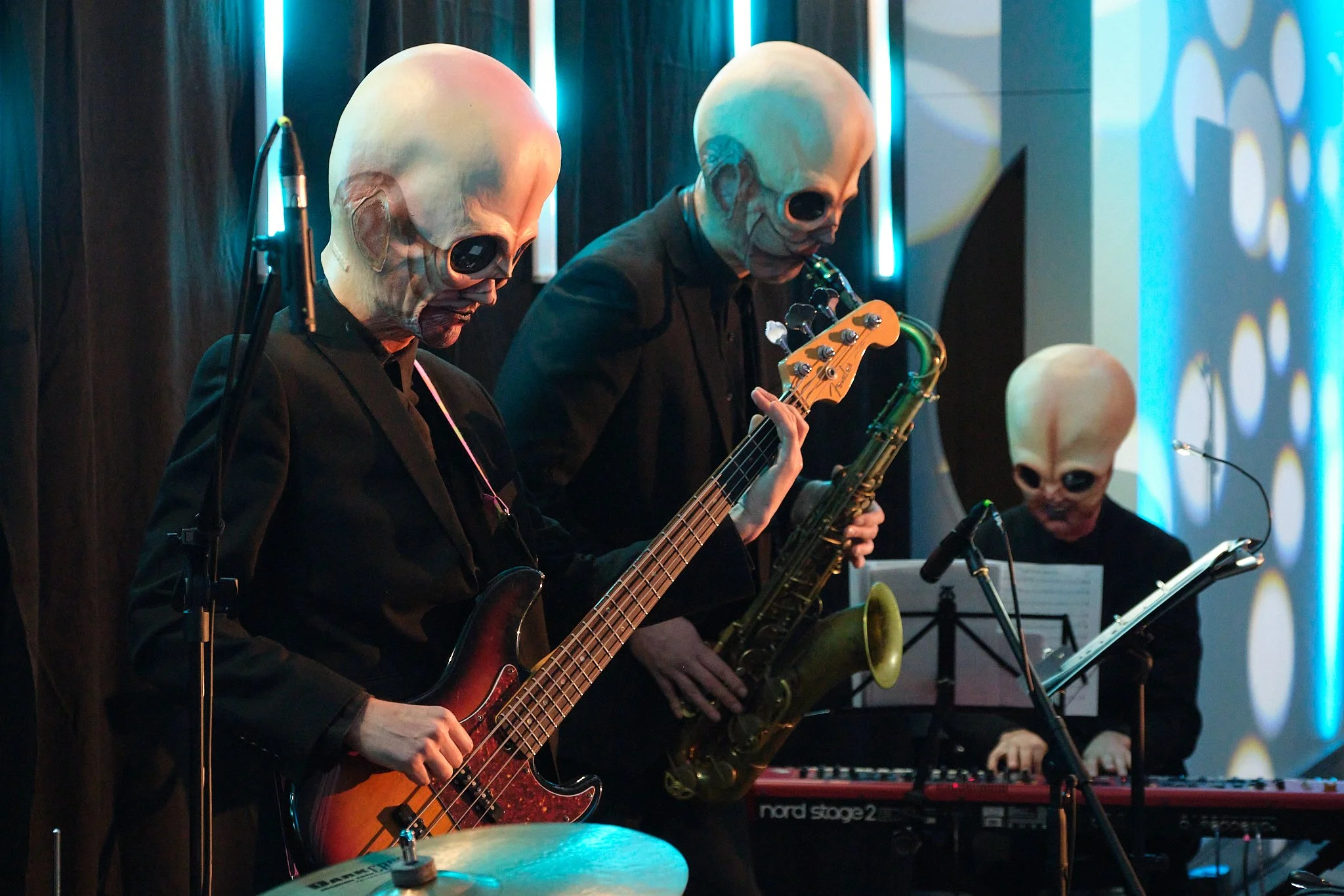 Three musicians in black suits and large bald skull masks perform on stage with various instruments, including electric bass, electric guitar, and saxophone, with blue neon lighting and patterned wall in the background. Aliens. Sci-fi cantina band.