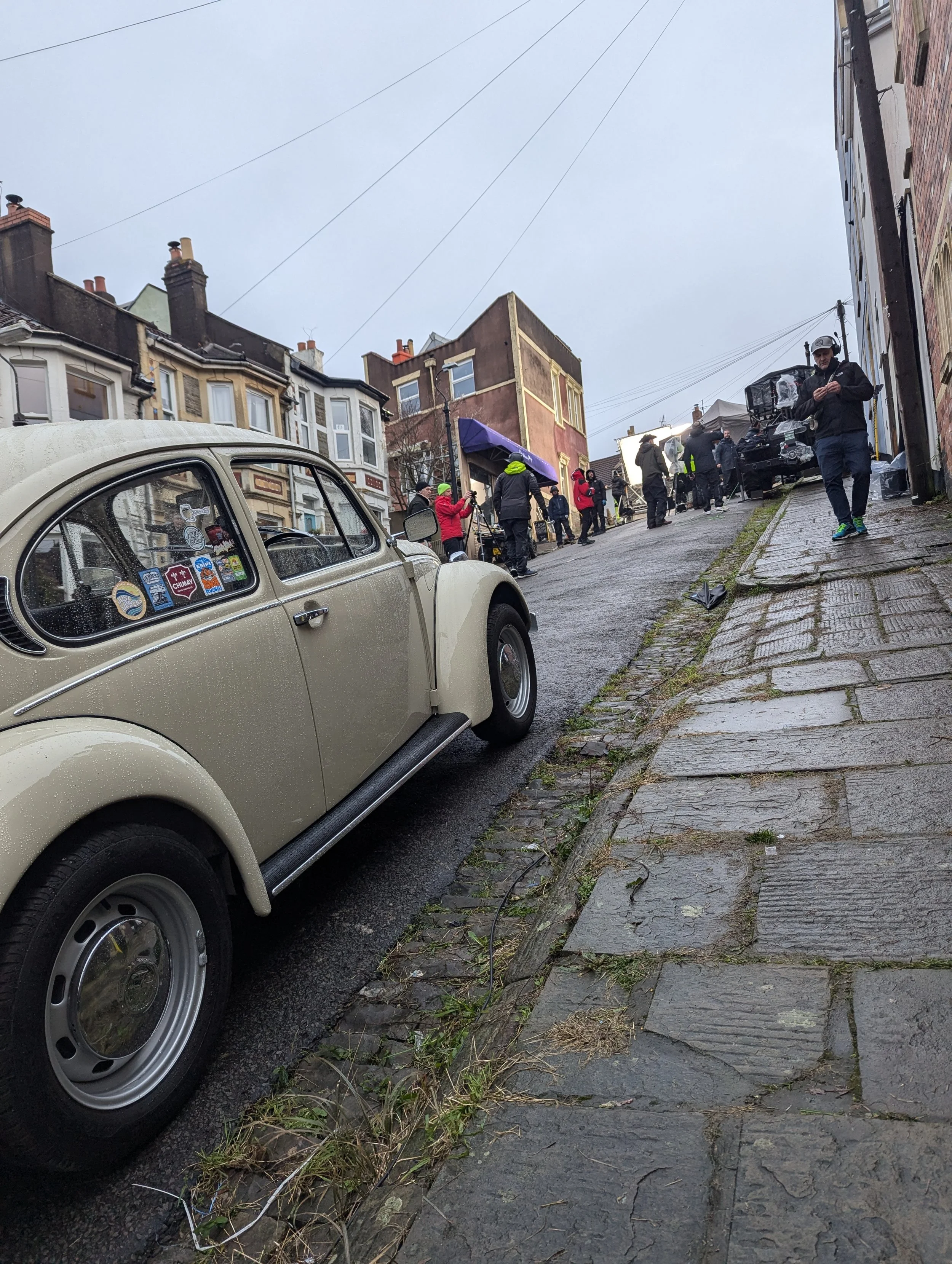 Street scene with vintage cream-colored car parked on wet street, people gathered under umbrellas and rainwear, film crew filming on a rainy day in an urban neighborhood with old buildings.