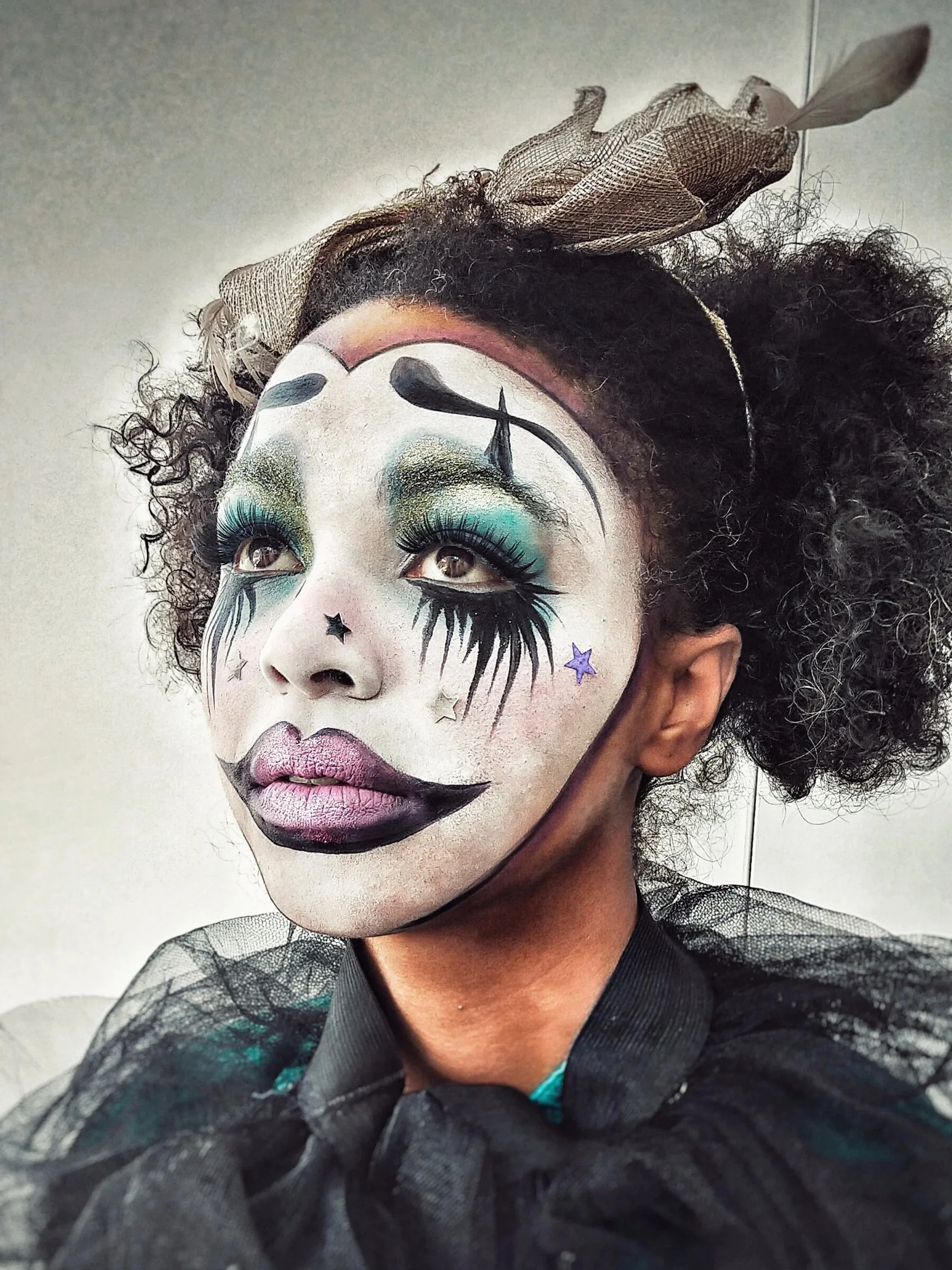 A person with clown makeup featuring white face paint, exaggerated black eyebrows, colorful eyeshadow, and black teardrop designs. They are wearing a black outfit with a bow tie and a decorative headpiece with tulle fabric, curly hair, and a headband.