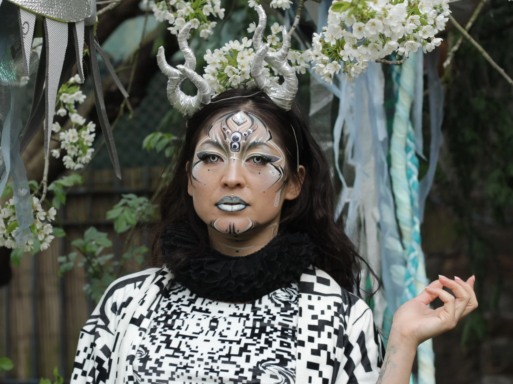 Woman with elaborate face paint resembling a mystical or fantasy creature, wearing a headdress with silver horns, surrounded by white flowers and blue and white ribbons.