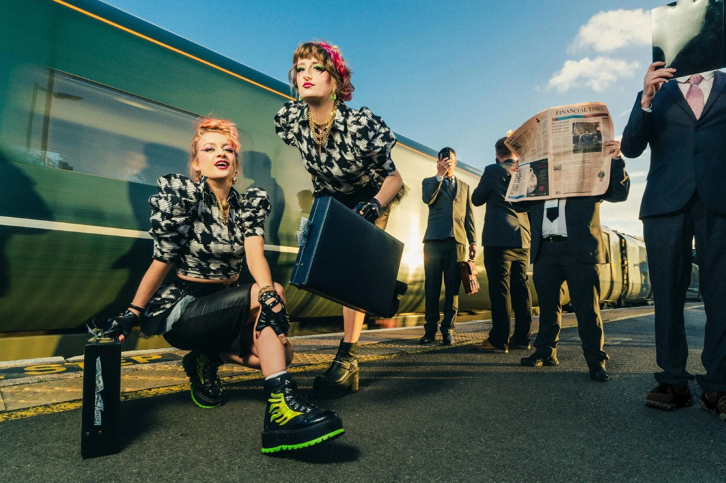 Two women dressed in punk fashion with brightly colored hair and bold makeup, waiting at a train station platform, with one squatting and the other standing, while several men in suits are in the background, some reading newspapers and taking photos.