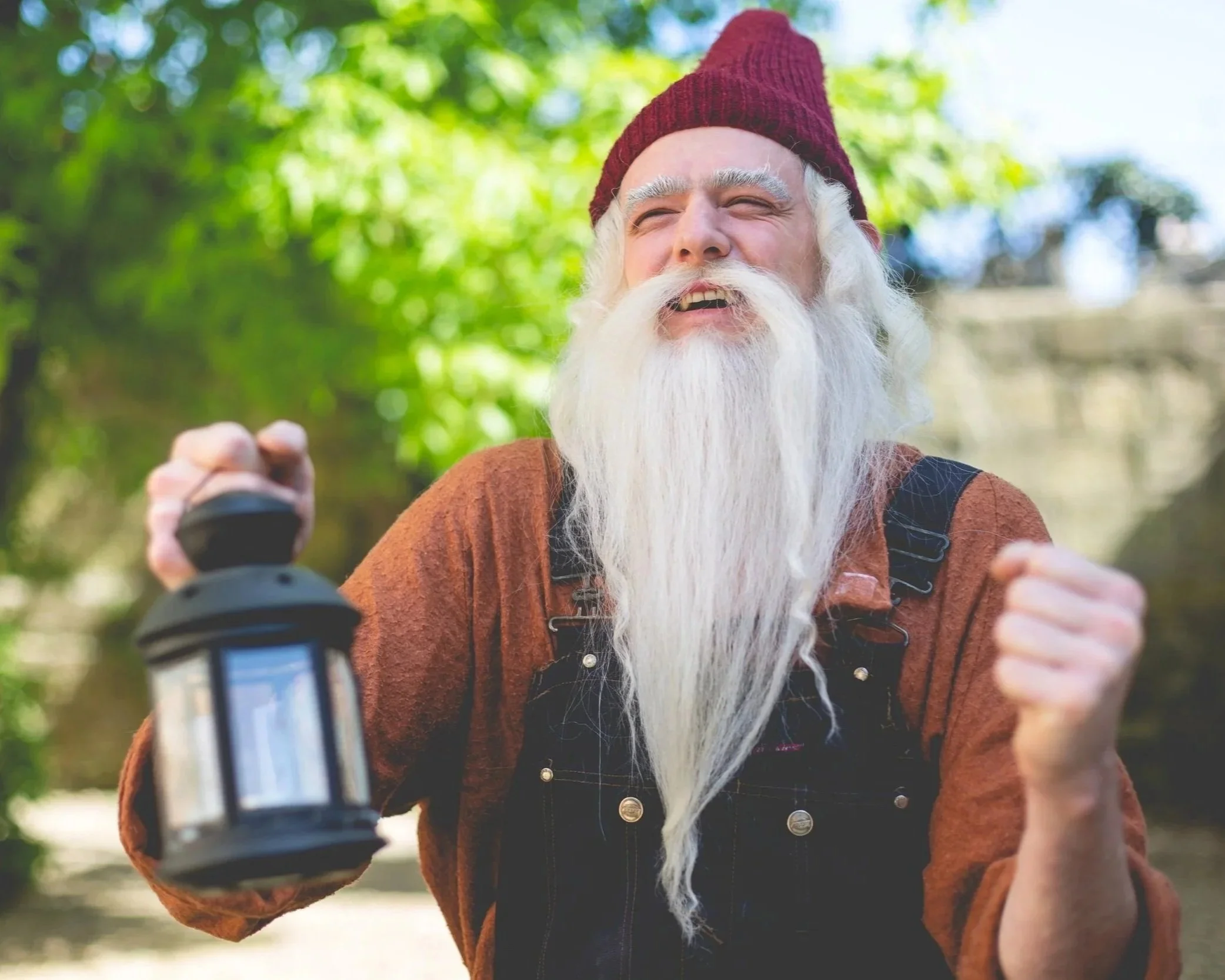 An elderly man with a long white beard and mustache, wearing a red beanie and brown shirt, holding a lantern in one hand and raising his fist in an outdoor setting with green trees in the background.