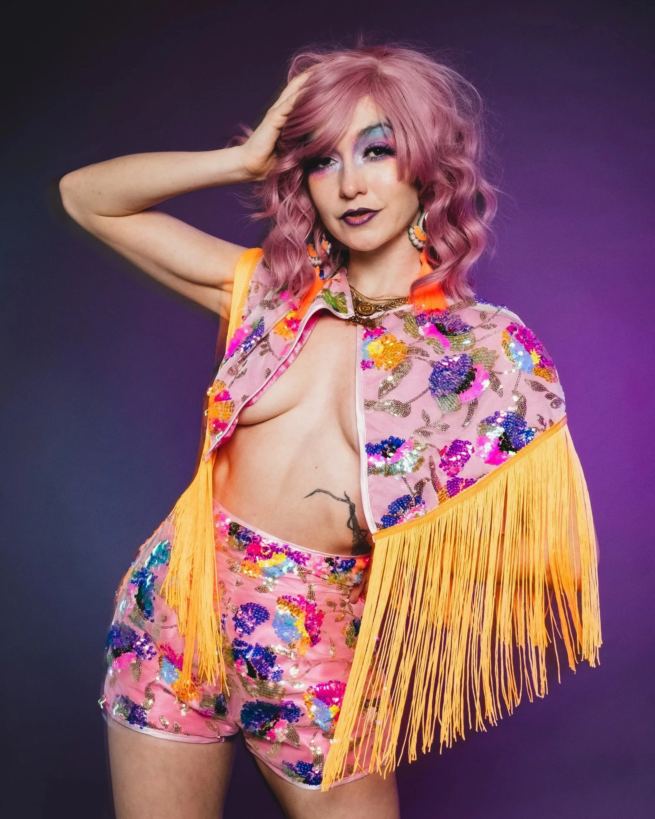 Woman with pink curly hair wearing a colorful floral jacket with fringes, matching shorts, and bold makeup, posing with one hand on her head against a purple background.