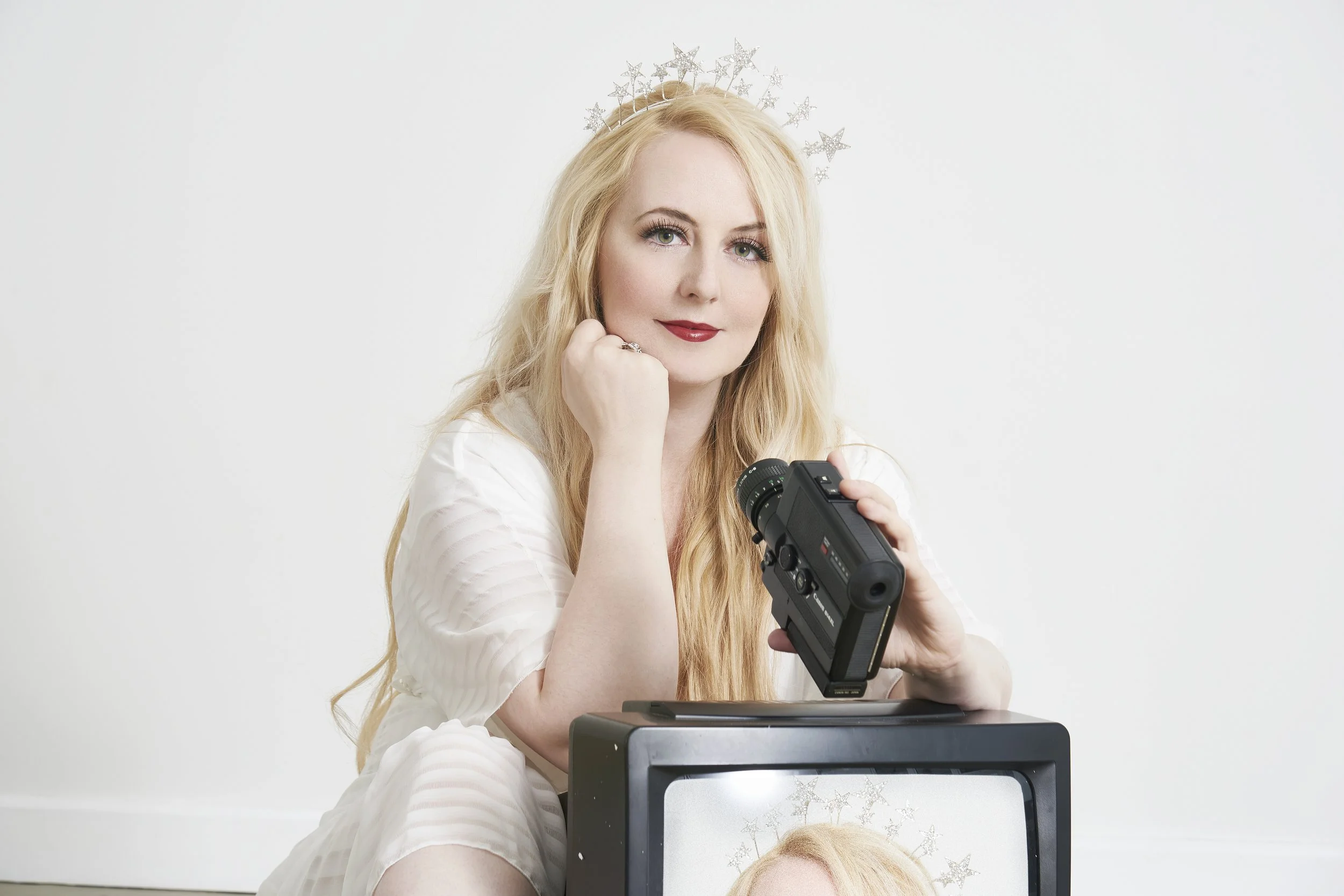 A woman with long blonde hair and a star crown, holding a video camera, sitting next to a TV showing her reflection.