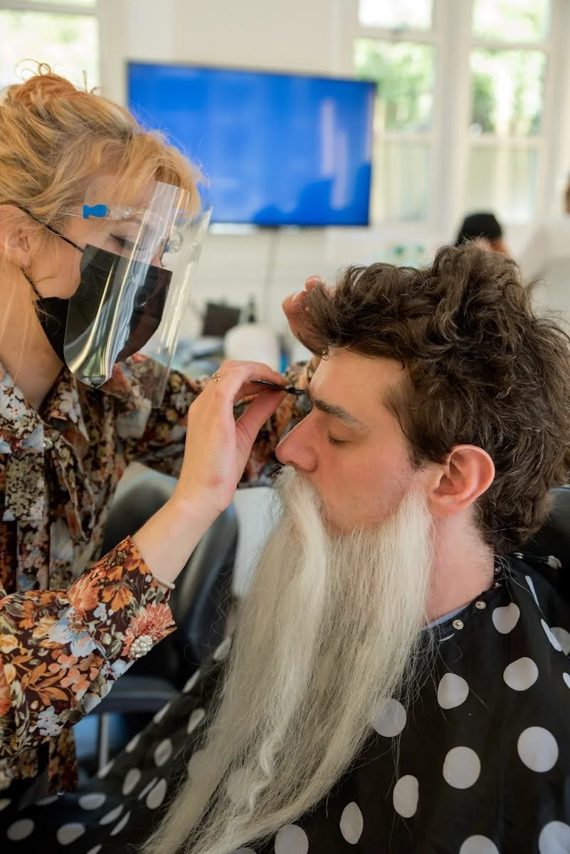 A woman wearing a face shield and a black mask is applying makeup or grooming a man's eyebrows. The man has a long white beard, curly hair, and is wearing a black smock with white polka dots. They are indoors in a bright room with large windows, a television in the background, and other people faintly visible.