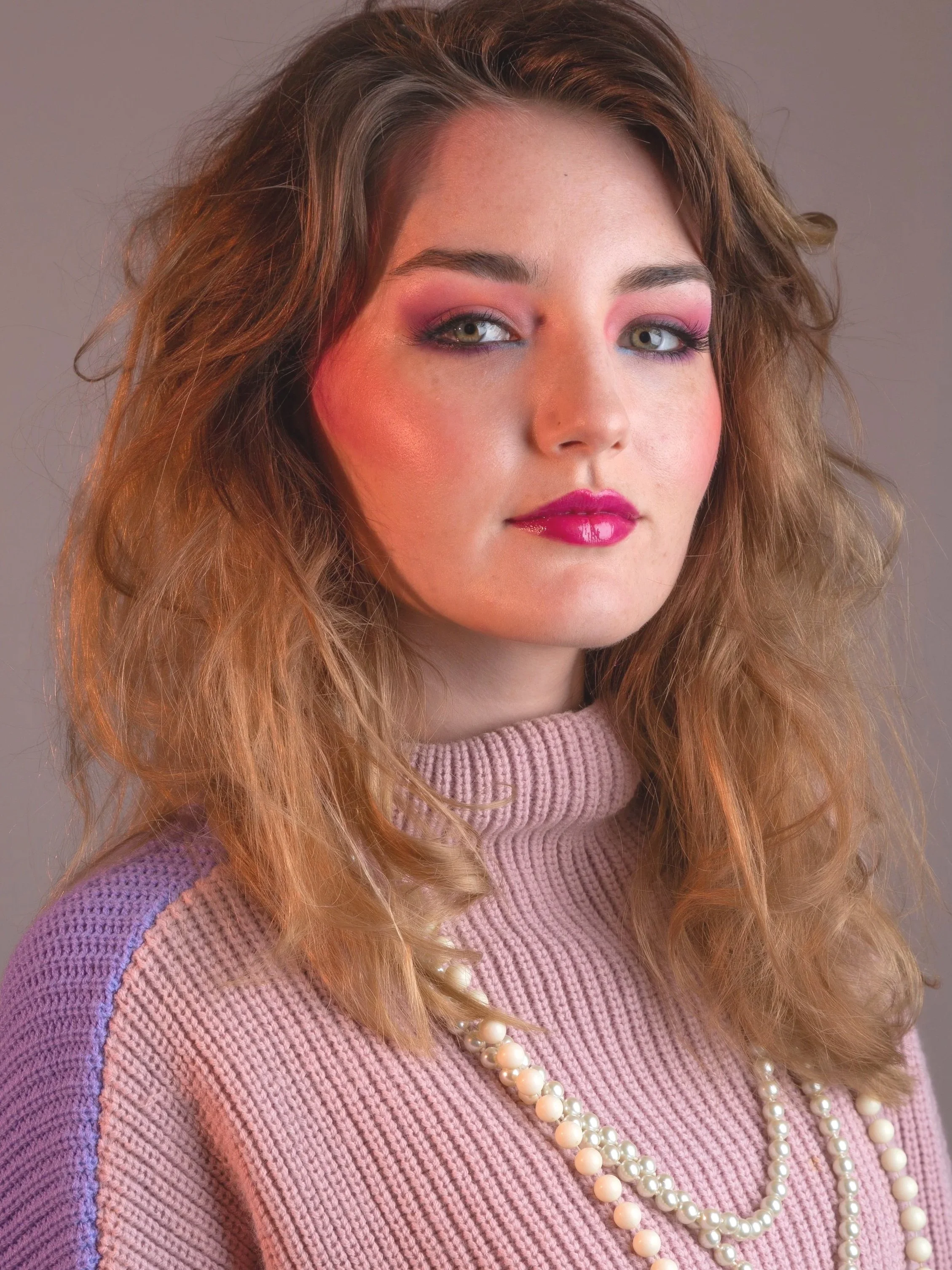 Close-up portrait of a young woman with shoulder-length, wavy auburn hair, wearing a pink knit sweater and multiple strings of pearls, with pink and purple eyeshadow and glossy pink lipstick, looking confidently at the camera.