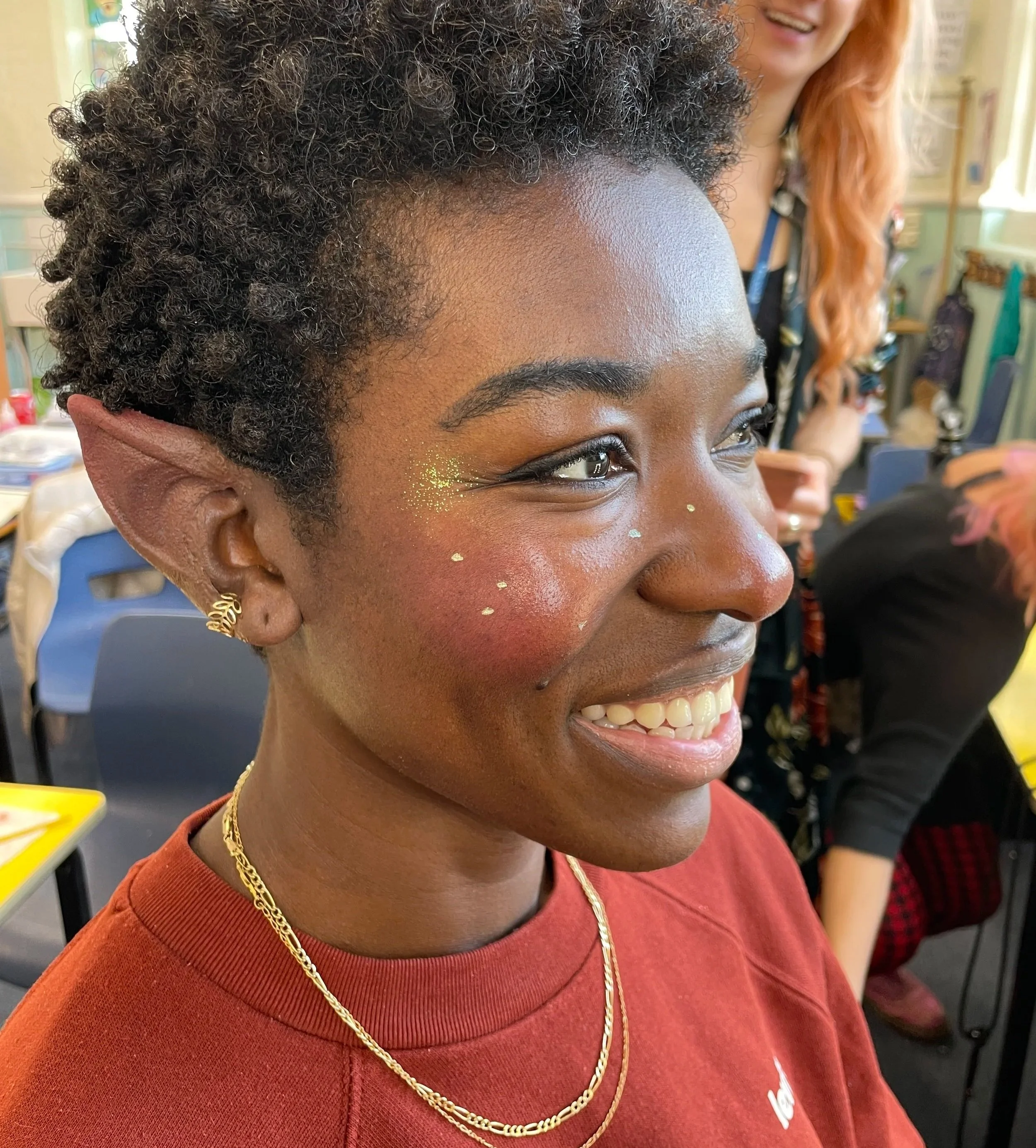 A person with curly short hair, elf ears, and gold jewelry, smiling with glitter on their face, wearing a red top in a lively indoor setting.