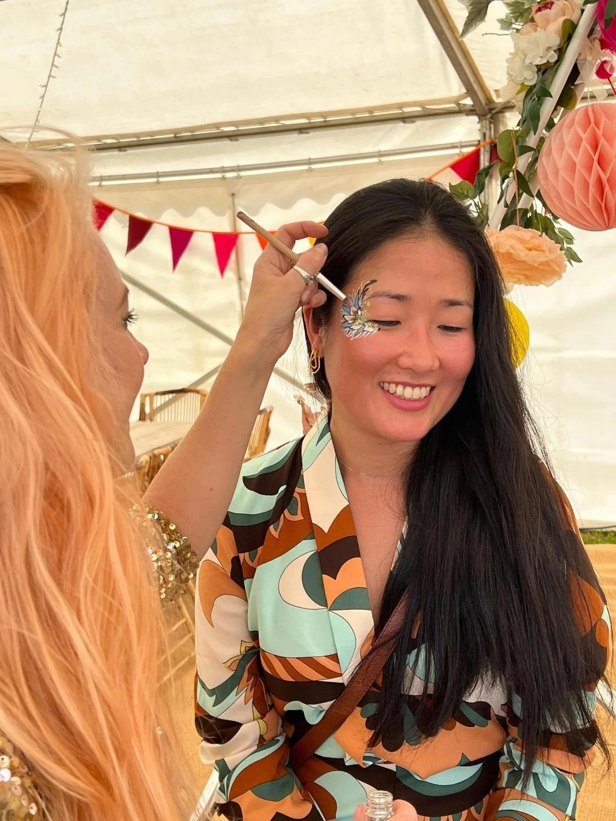 A woman sitting and smiling while another woman paints a colorful, intricate design on her face at an outdoor event under a tent decorated with pink, peach, and yellow paper lanterns and bunting.