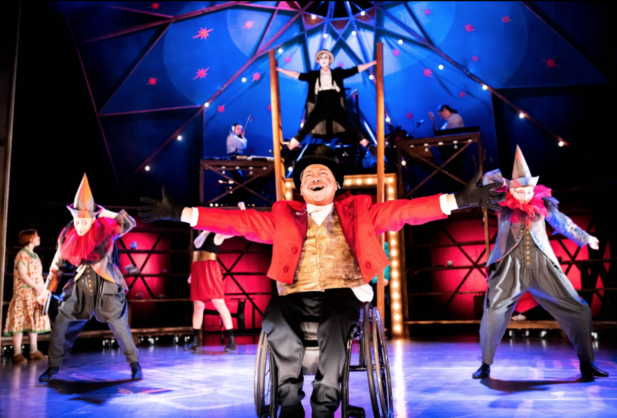 Circus performers on stage, including a man in a wheelchair with arms outstretched, smiling, flanked by two clown characters, with acrobats in the background