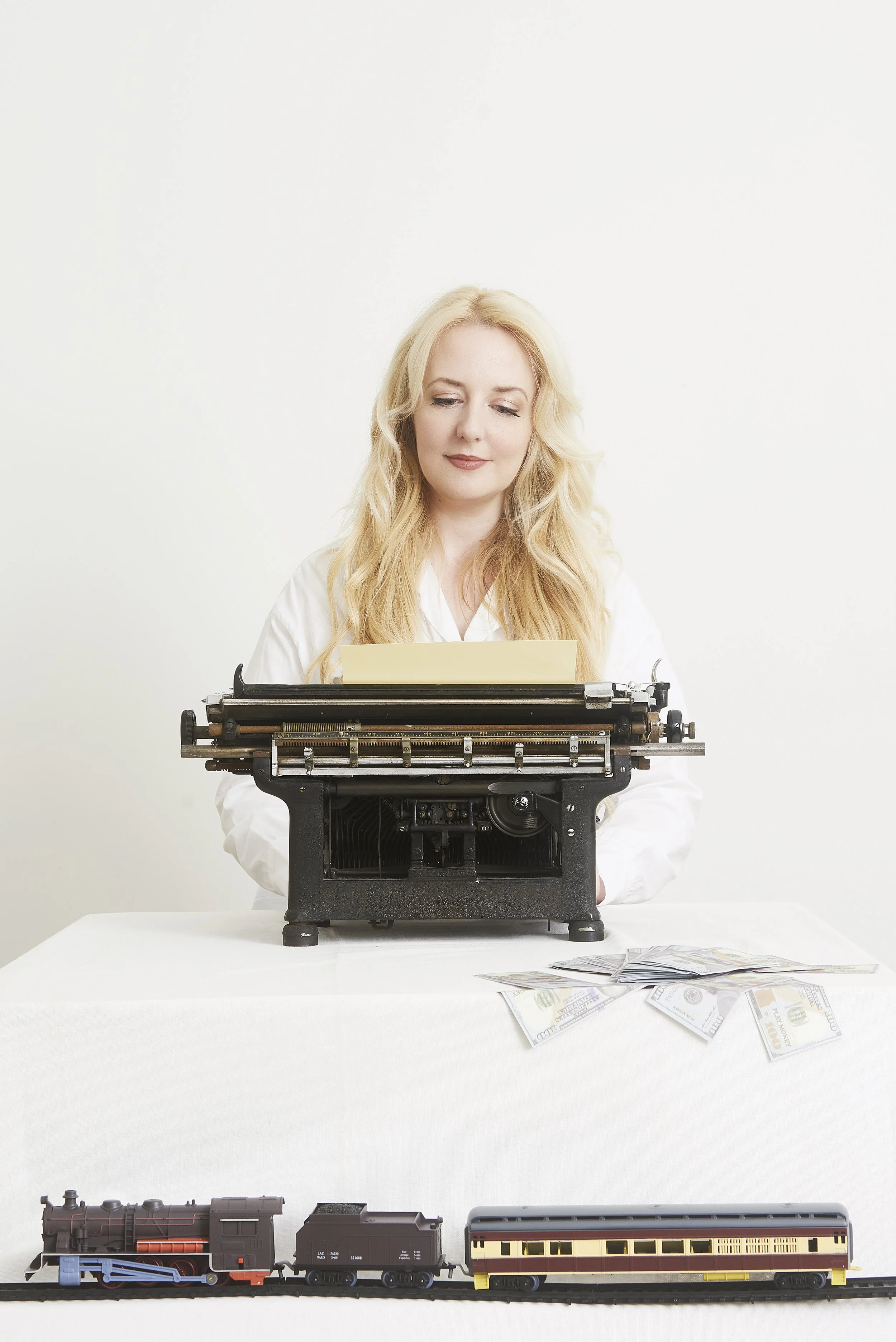A woman with blonde hair seated at a table using a vintage typewriter, with scattered fake U.S. currency bills and a toy train on a track in front of her, against a plain white background.