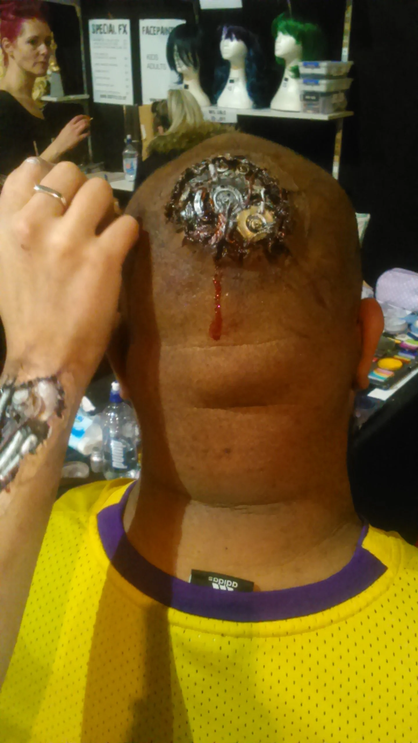 A person with a shaved head, wearing a yellow sports jersey, has a realistic artificial injury on their scalp with a wound, exposed tissue, and blood.