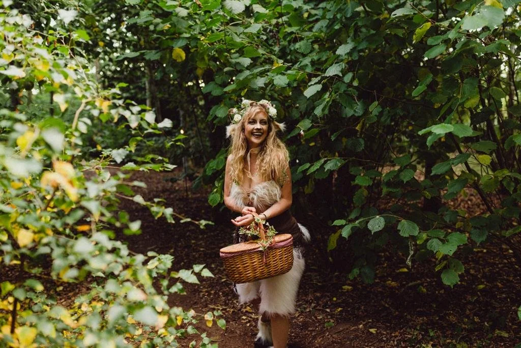 A woman with long, curly blonde hair dressed in a whimsical costume with a floral crown, holding a wicker basket, standing in a lush green forest.