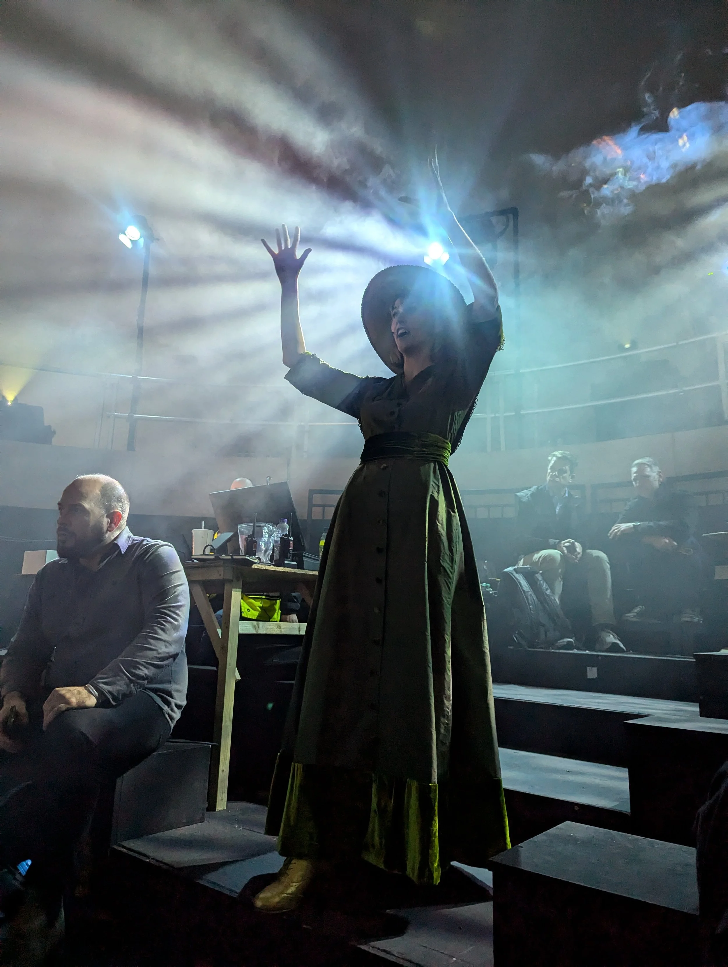 A woman dressed in a vintage long dress and wide-brimmed hat is dancing on a stage with dramatic lighting, surrounded by an audience.