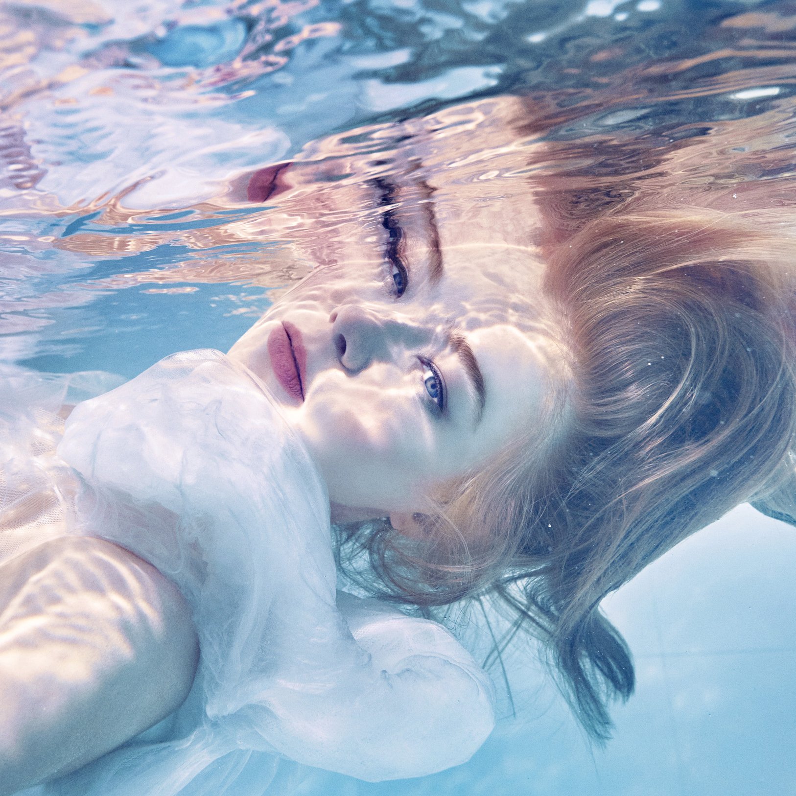 A woman lying in a pool with her face partially submerged in water, her eyes open and looking upward, with sunlight casting patterns on her face and water around her.