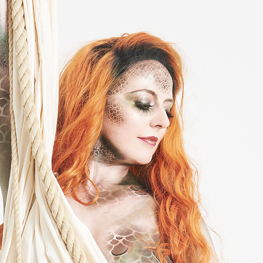 A woman with long red hair displaying scales and a textured pattern on her face and body, with her eyes closed, standing beside a cream-colored curtain.