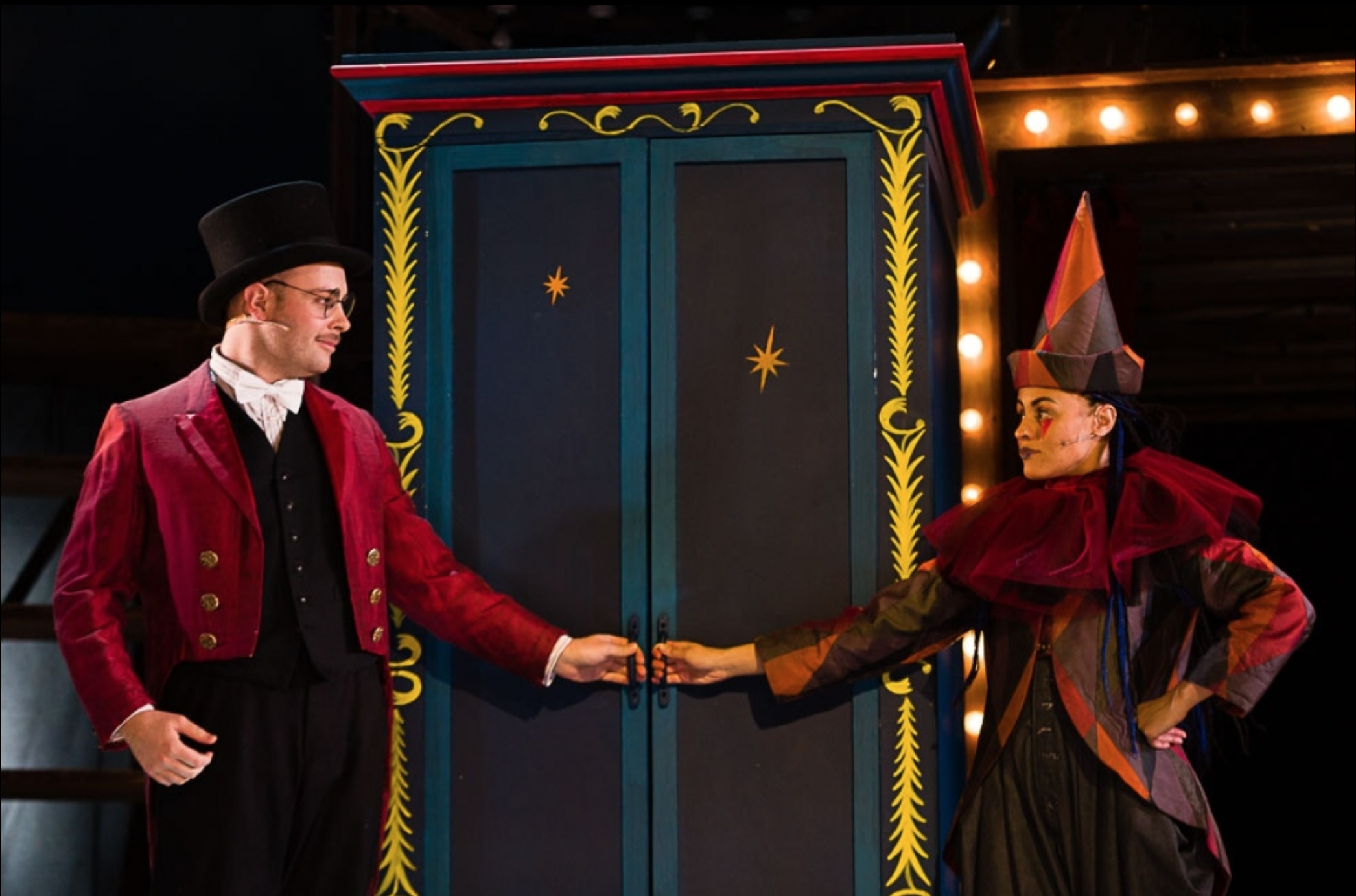 Two performers in colorful costumes connect hands in front of a decorative door on stage. One wears a top hat and vintage attire, the other a tall, striped cone hat and a ruffled, multi-colored outfit. The background includes warm stage lighting.
