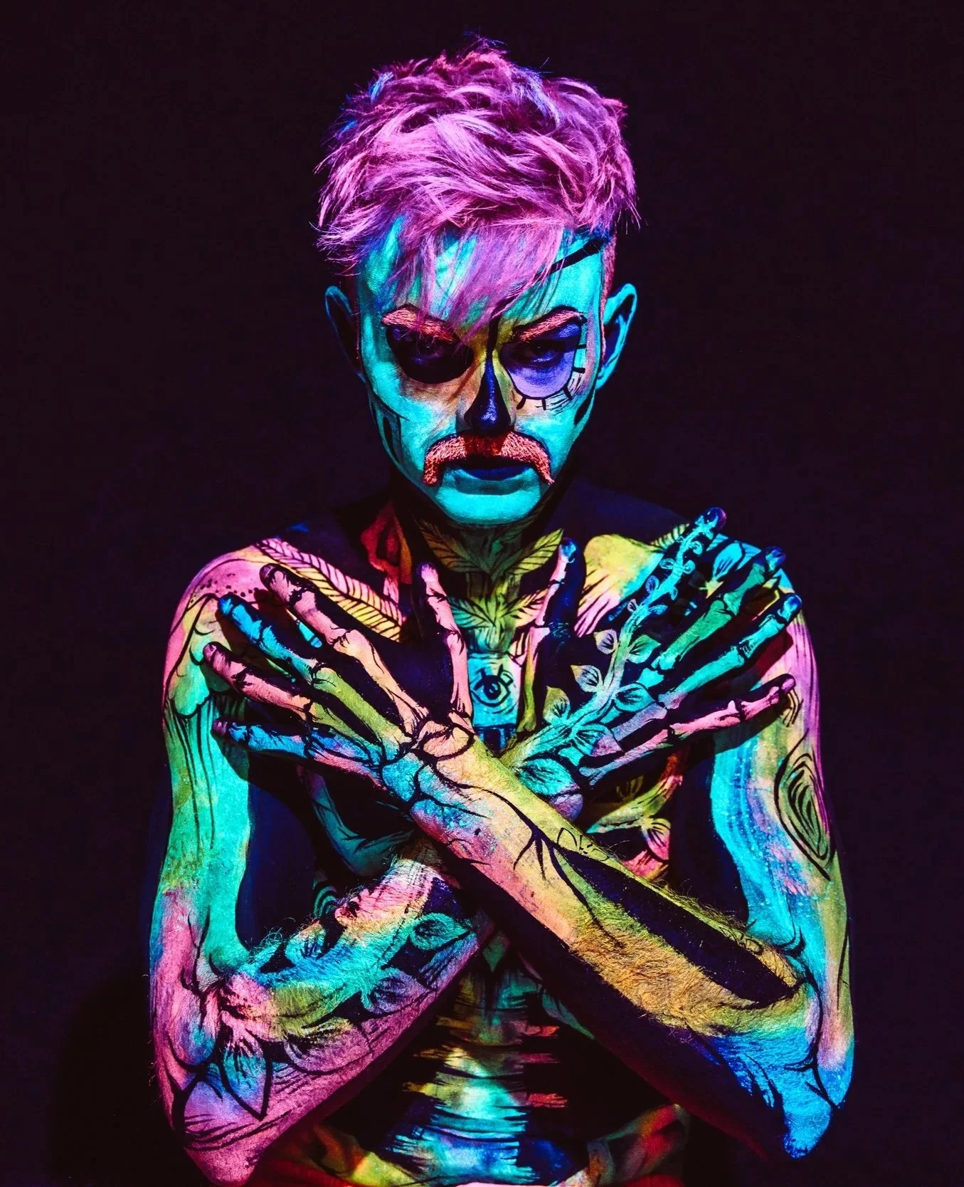 A person with neon face and body paint, styled like a skeleton, with colorful tattoos on their arms, crossing their arms over their chest, against a dark background.