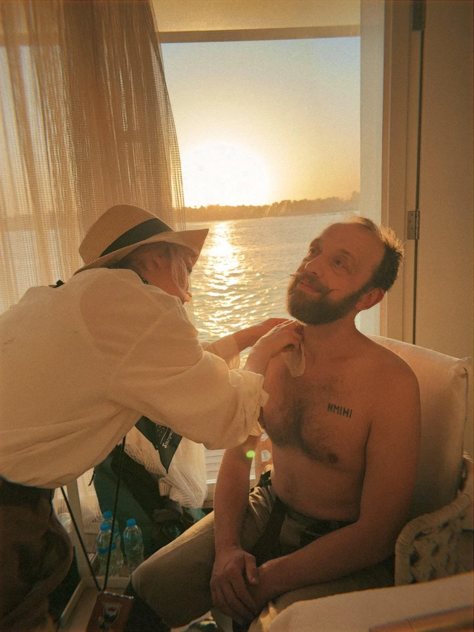 A woman in a sunhat and white blouse is applying makeup or face paint to a shirtless man with a beard, sitting on a chair by a large window showing a sunset over a body of water.