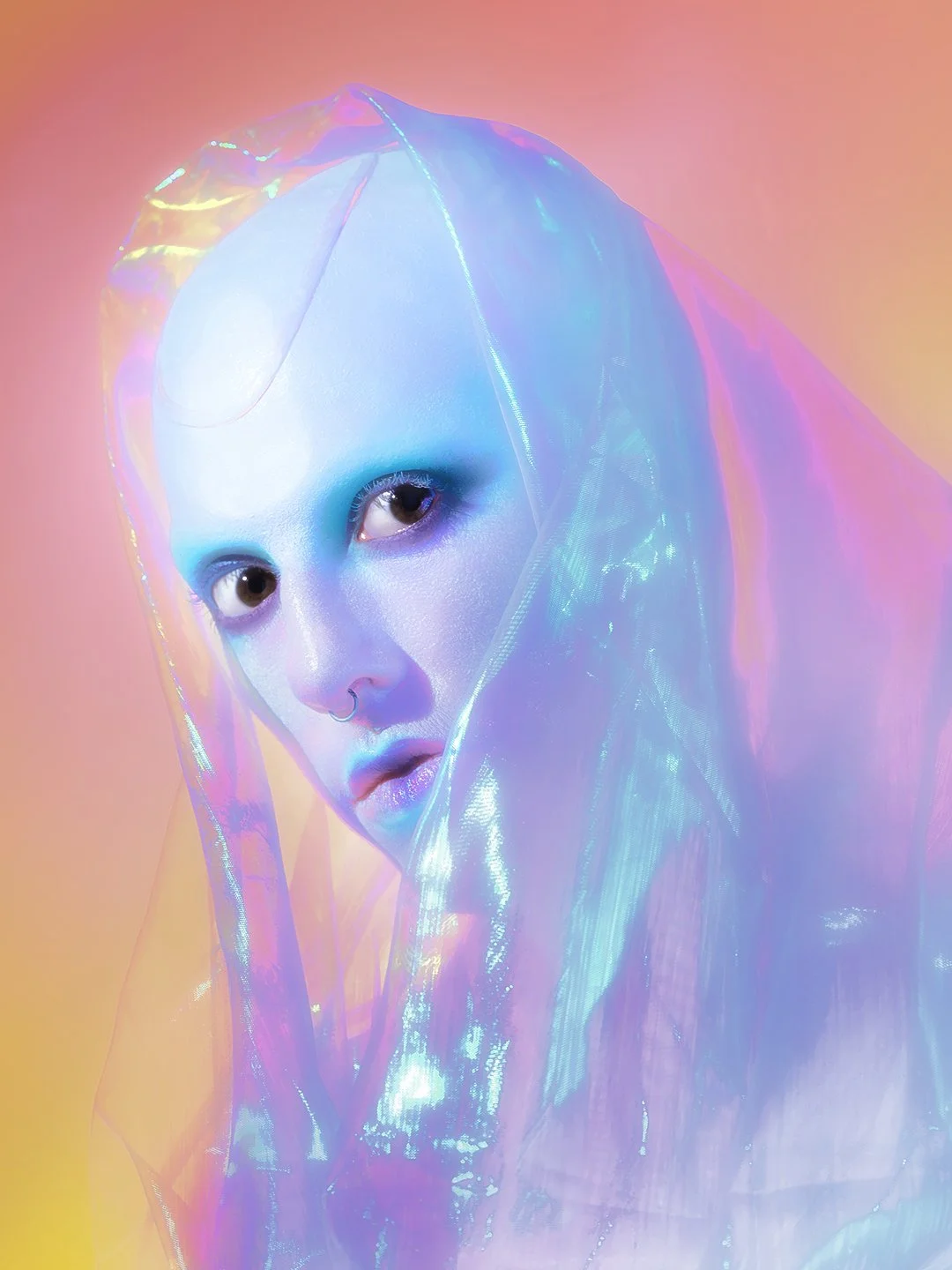 A woman with an iridescent holographic veil and blue makeup, looking at the camera against a gradient pink and orange background.