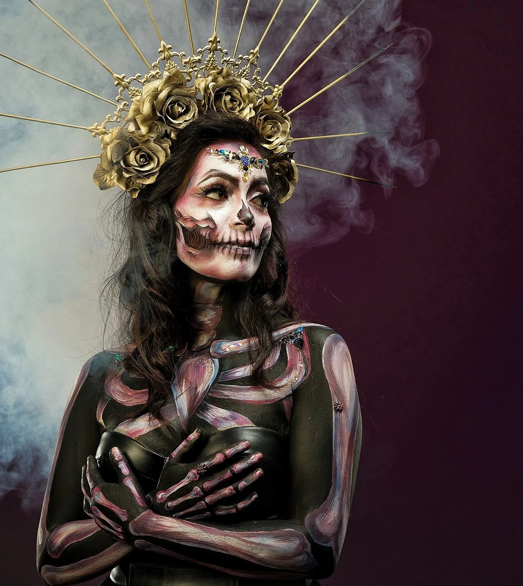 A woman with face and body painted like a skeleton, wearing a gold floral crown and a halo-like headpiece with roses and radiating rods, standing against a dark, smoky background.