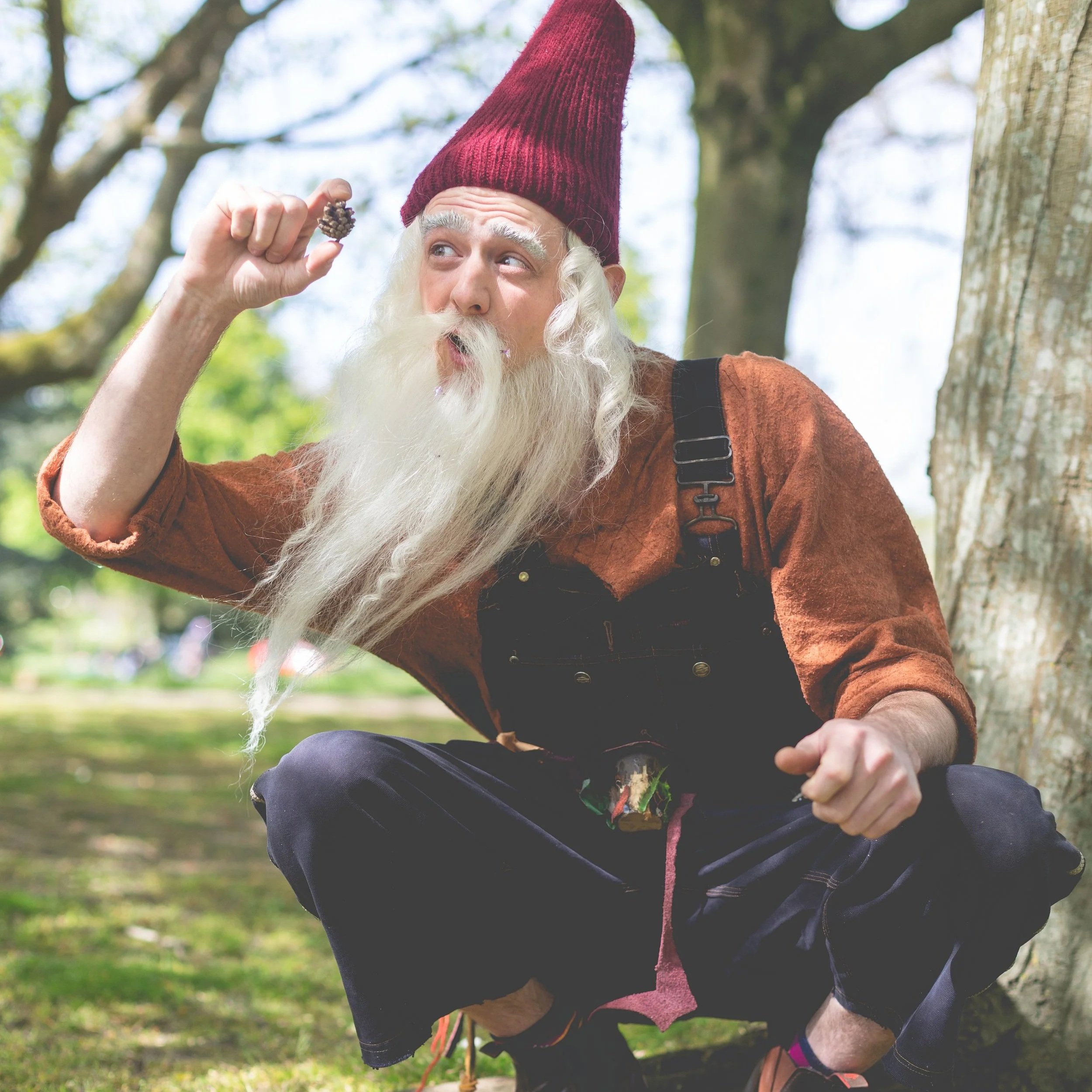 An elderly man with a long white beard and hair, wearing a red pointed hat, orange shirt, and black overalls, sitting outdoors in a park with trees. He is holding a pine cone and appears to be examining or about to eat it.