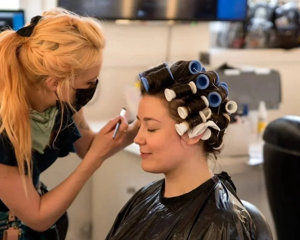 A woman with rolled hair in curlers getting a makeup application from a makeup artist in a salon