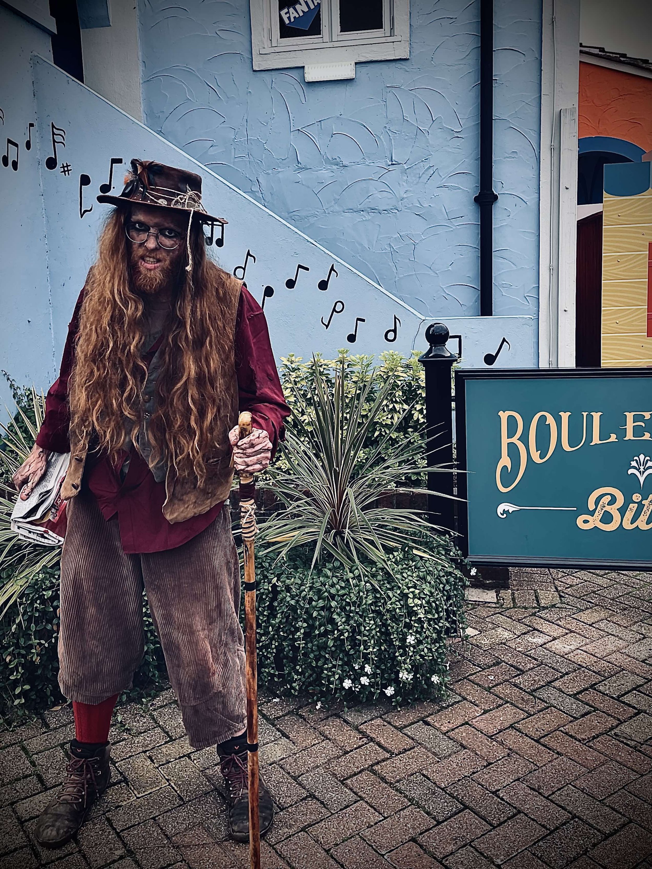 A person dressed as a wizard or wizard-like character with long curly hair and beard, wearing a hat and glasses, standing outdoors near a sign that reads 'Boule B.' The person is holding a staff and has a mischievous expression. Background includes a wall with musical notes and a window.
