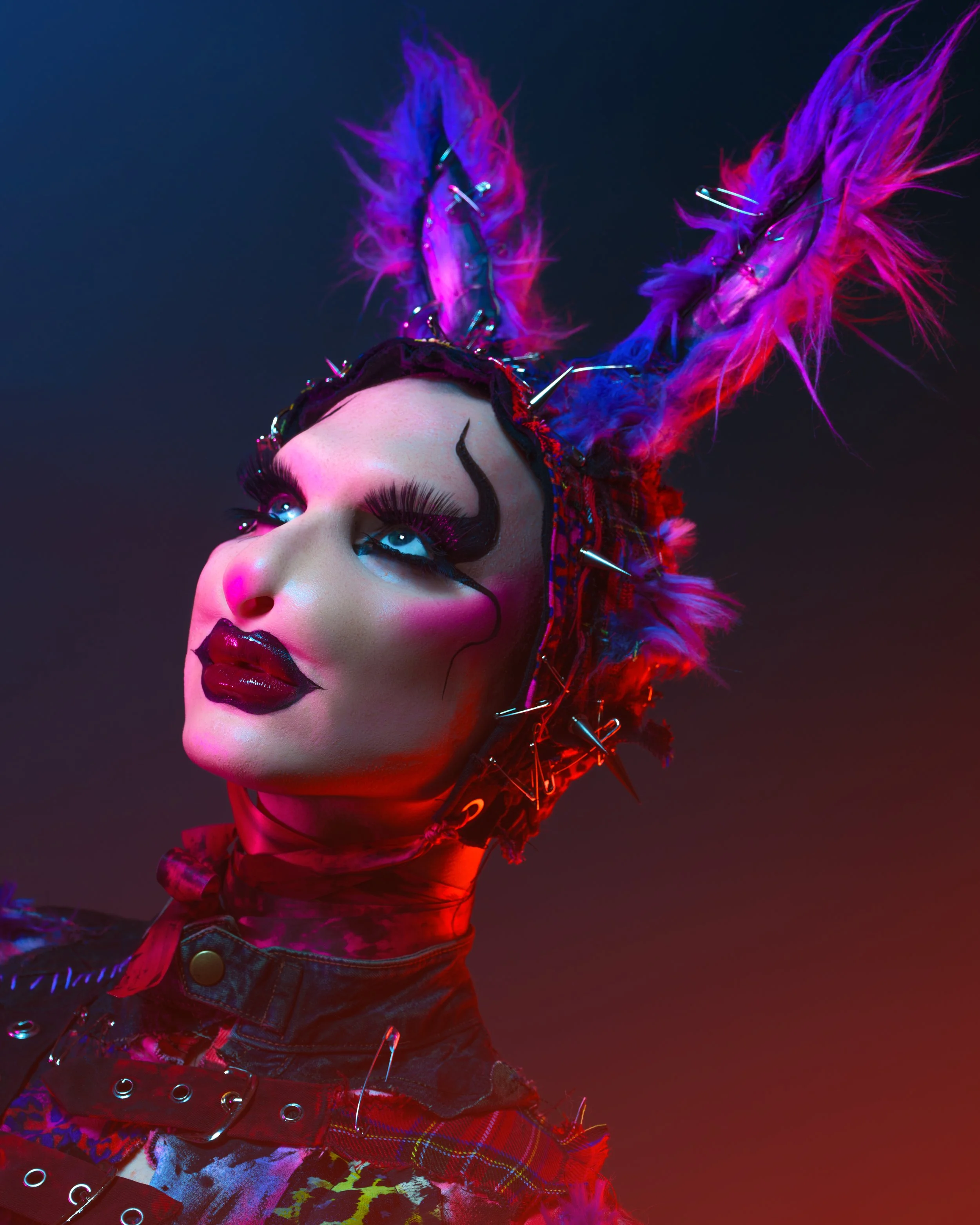 A person with elaborate makeup and costume, including bright blue eyes, dark lipstick, and dramatic eyelashes, wearing a headpiece decorated with colorful feathers, spikes, and fabric.