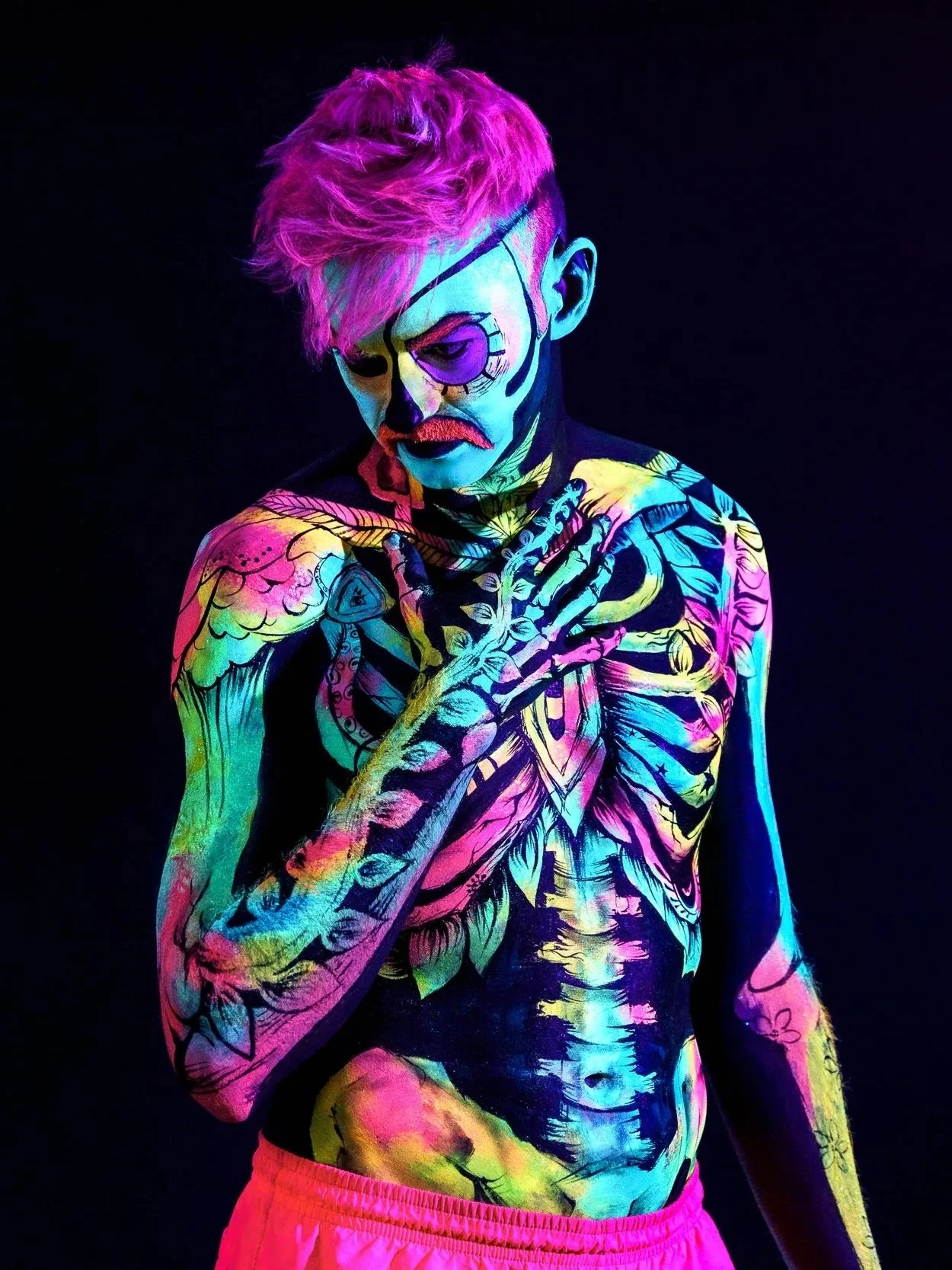 Person with body painted in colorful neon designs, including skeletal motifs, with pink hair, facial makeup resembling a skull, and wearing pink shorts against a black background.