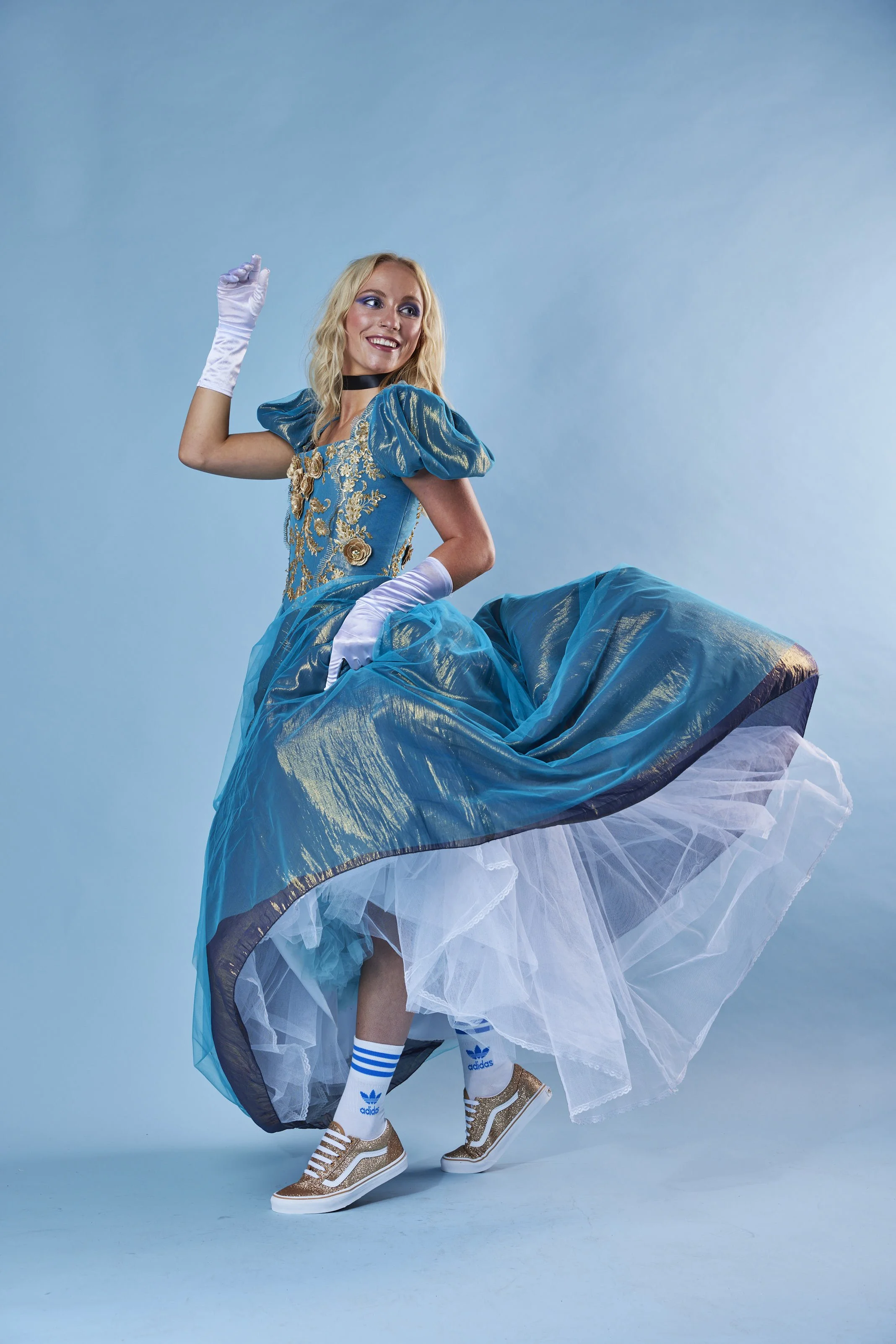 Woman dressed as Cinderella in a blue ball gown, white gloves, and gold sneakers, posing playfully on a blue background.
