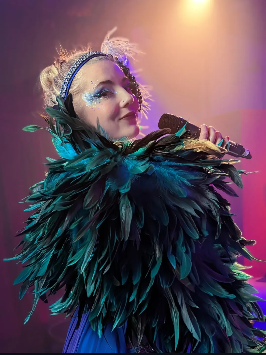 A performer with elaborate feathered costume, glitter makeup, and a tiara, holding a microphone on stage with colorful lighting.