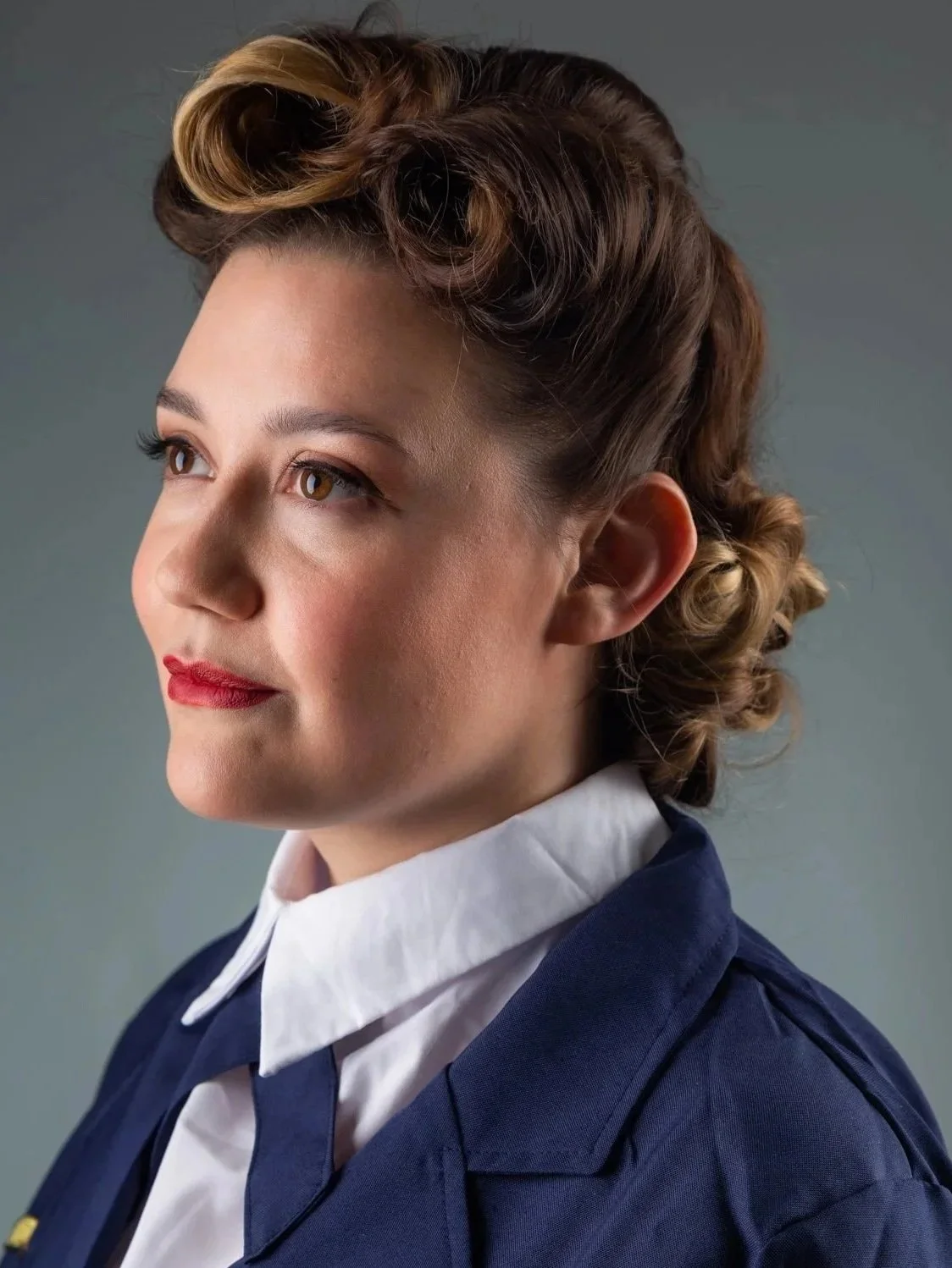 Portrait of a woman with vintage hairstyle, wearing a navy jacket and white collared shirt, against a gray background.