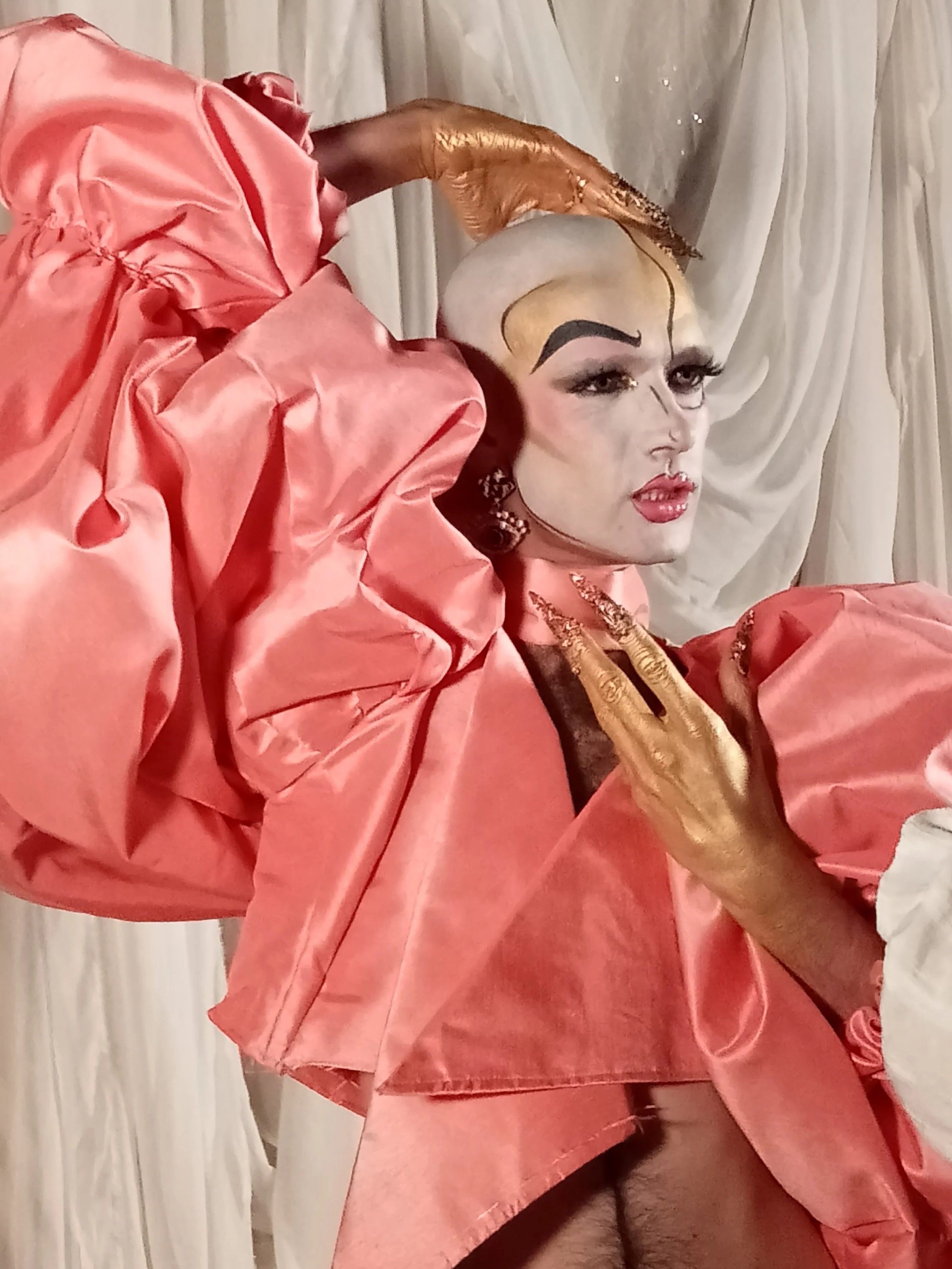 A person with dramatic makeup and a bold hairstyle wearing a shiny pink garment, striking a pose with one hand on their chest and head tilted back, in front of a light-colored curtain.
