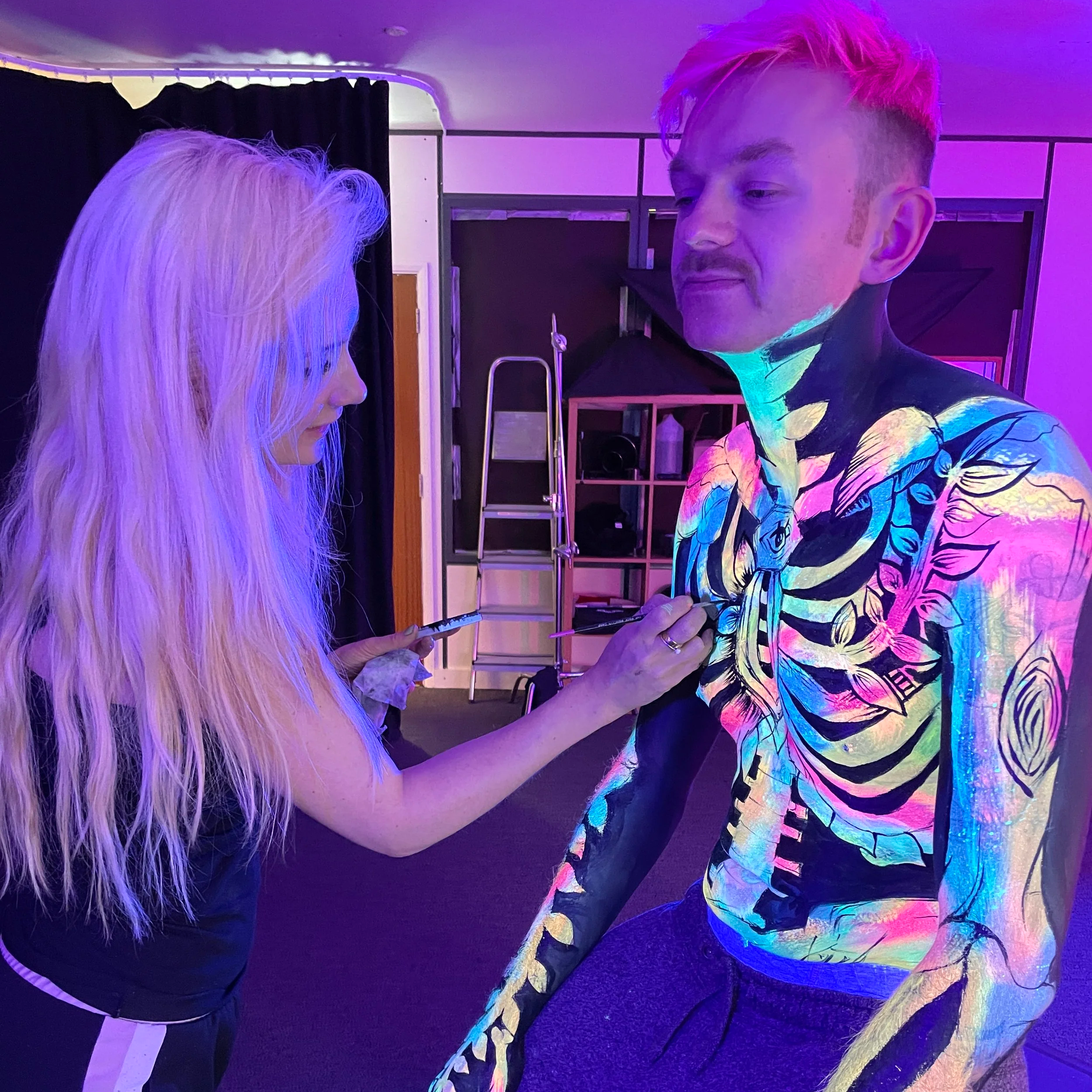 A woman paints bright, colorful designs on a man’s upper body and arm, with both under blacklight, revealing a vivid, glowing body art pattern.