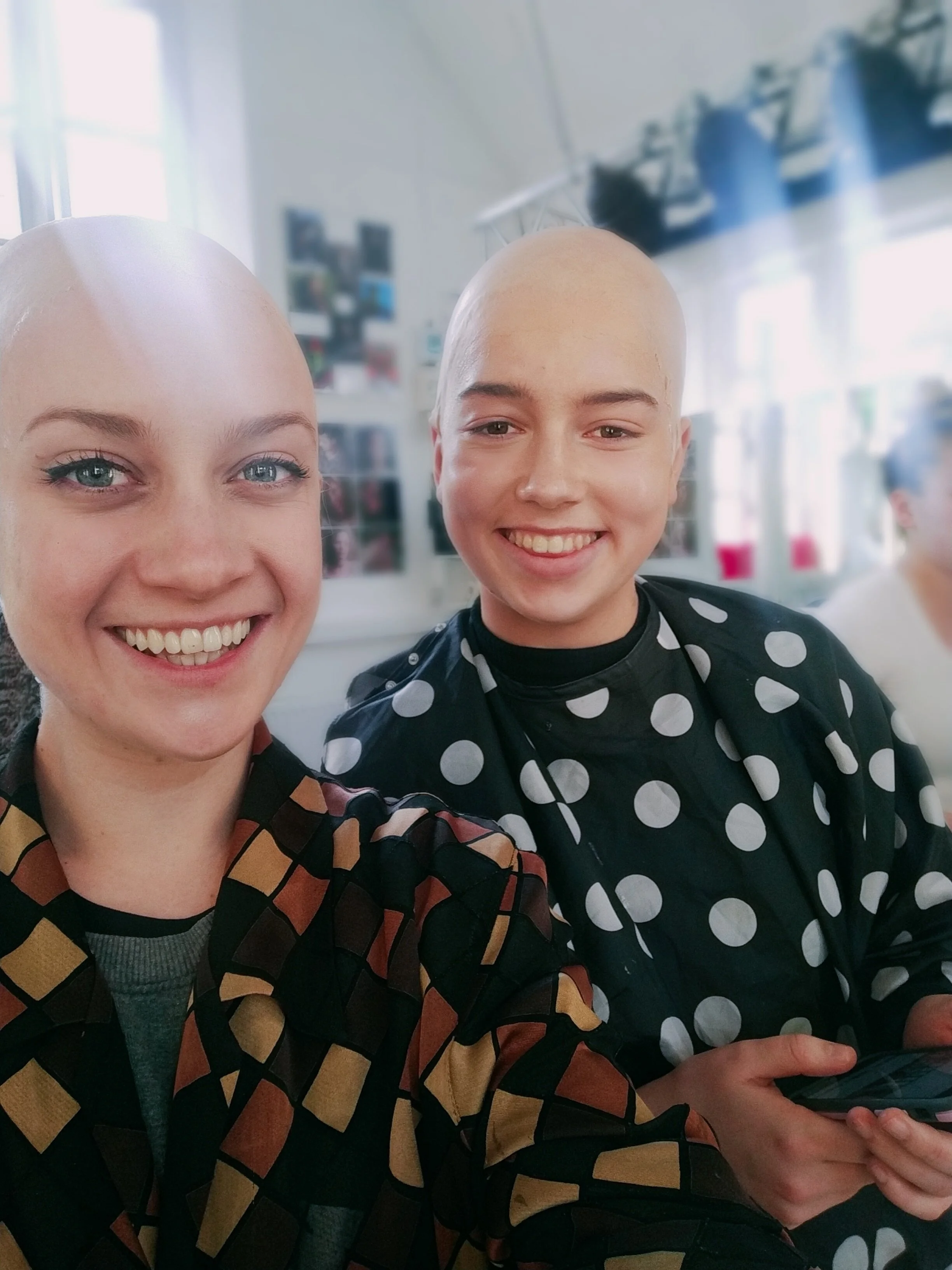 Two young women with shaved heads smiling at the camera, wearing patterned clothing, in a brightly lit room with photos and decorations on the wall behind them.