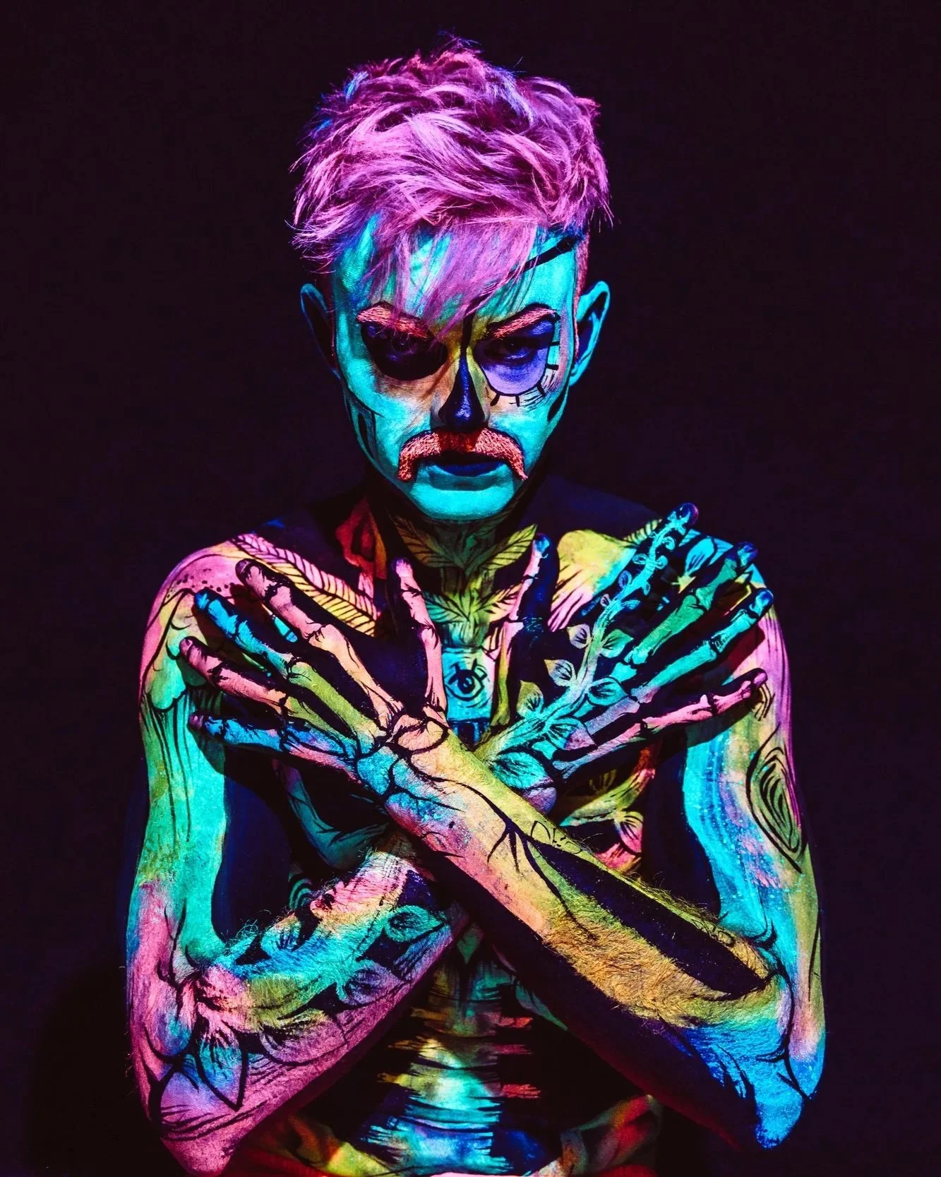 Person with body painted in vibrant neon colors, crossed arms, and face painted to resemble a skeleton and skull. Black background.
