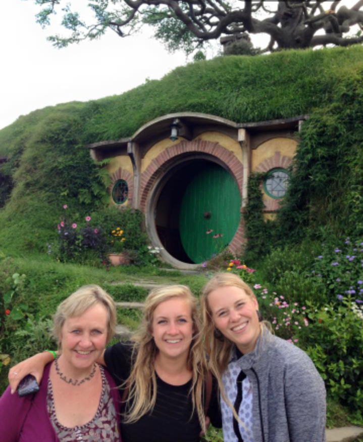 Three smiling women standing outdoors in front of the Hobbit house from Bilbo Bott's House, an earth-covered home with a round door, surrounded by colorful flowers and greenery.