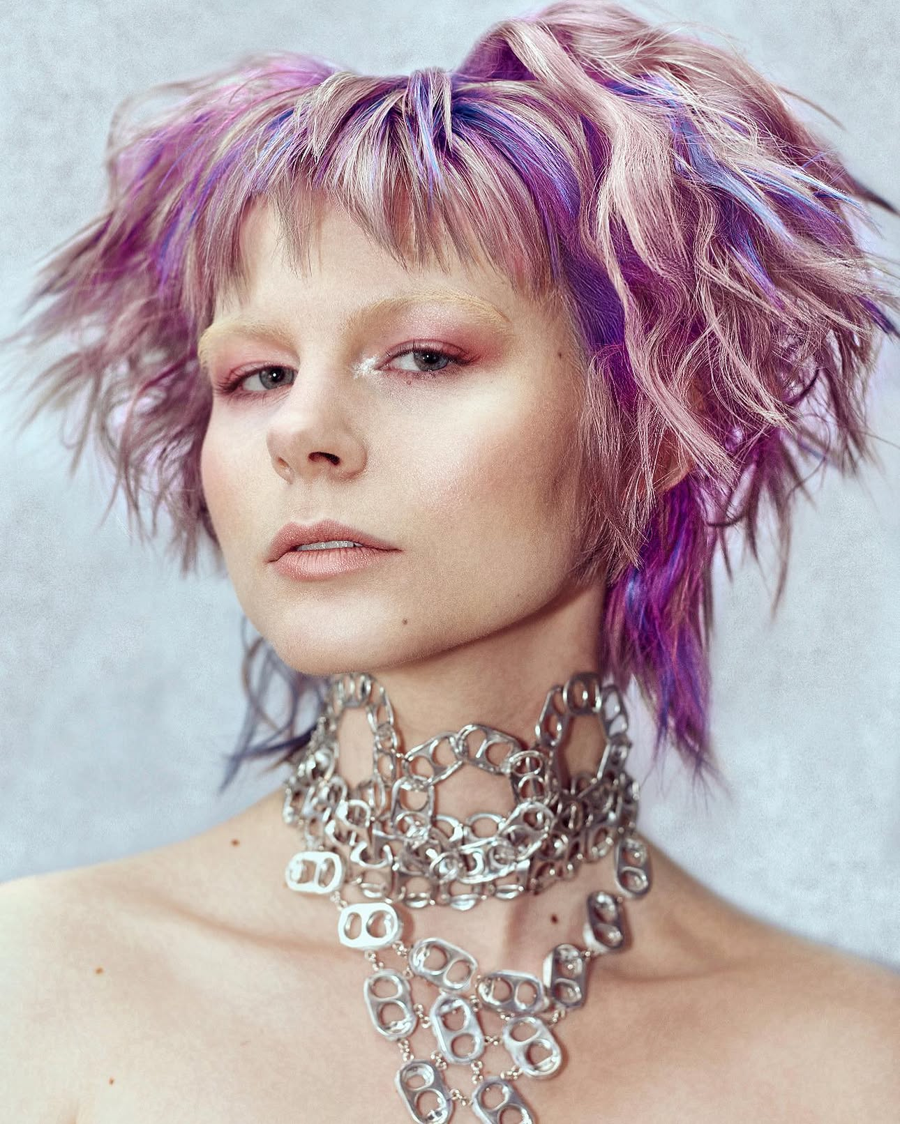 A woman with short, tousled hair dyed in pastel pink, purple, and blue shades, wearing multiple chunky silver chain necklaces.