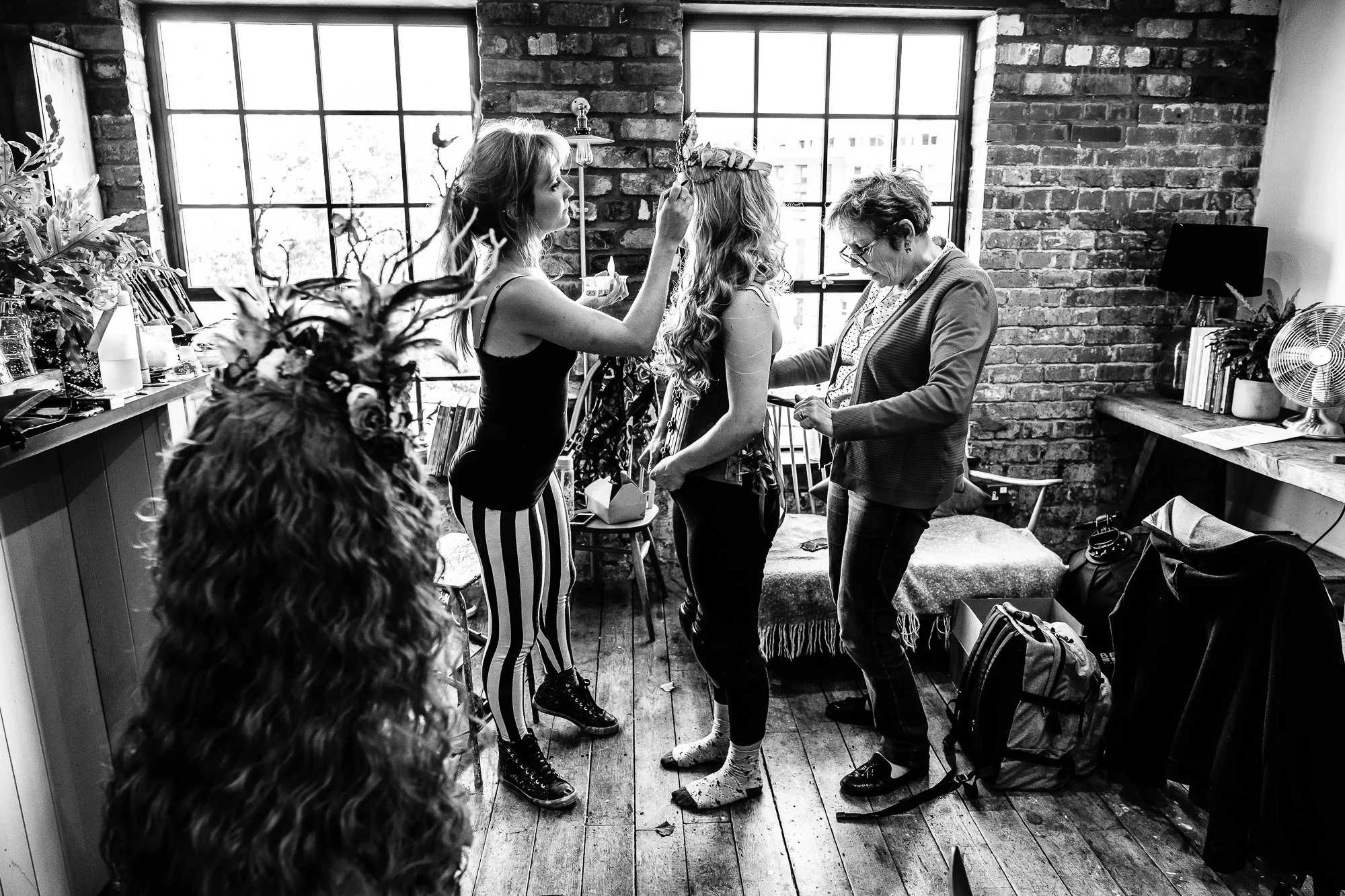 Two women, one with a crown, preparing for a wedding in a cozy brick-walled room with large windows. One woman is putting on the crown, while an older woman helps with the dress. A third woman with curly hair stands nearby, and the room is filled with personal items and books.