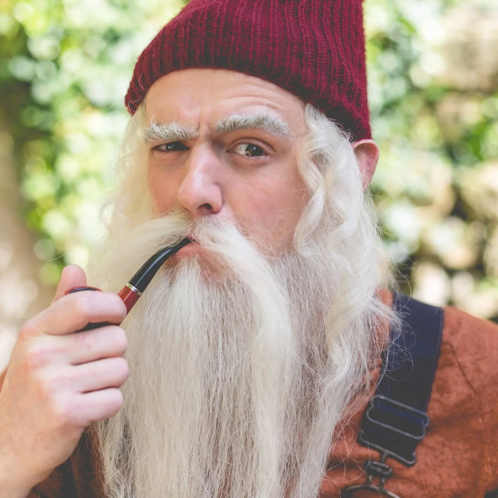 An elderly man with a long white beard and white eyebrows, wearing a maroon knit cap and a brown shirt, is holding a smoking pipe in his mouth and winking at the camera with a playful expression. The background is blurred with green foliage.
