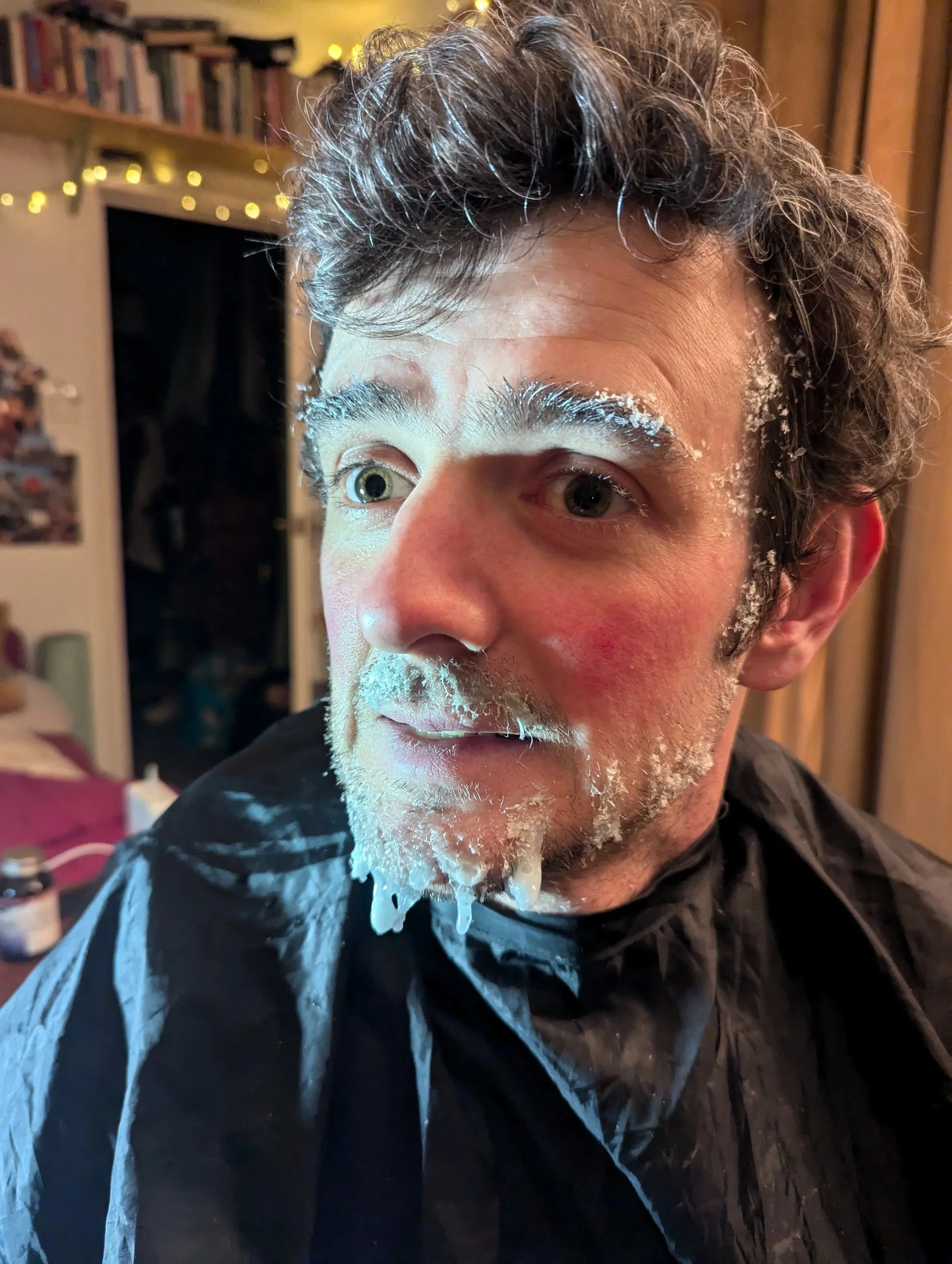 A person with messy hair and facial hair has a humorous mess of white cream or foam on their face, around the mouth, nose, and eyebrows. They are wearing a black barber cape, and the background includes a room with wooden paneling, books, and a closet.