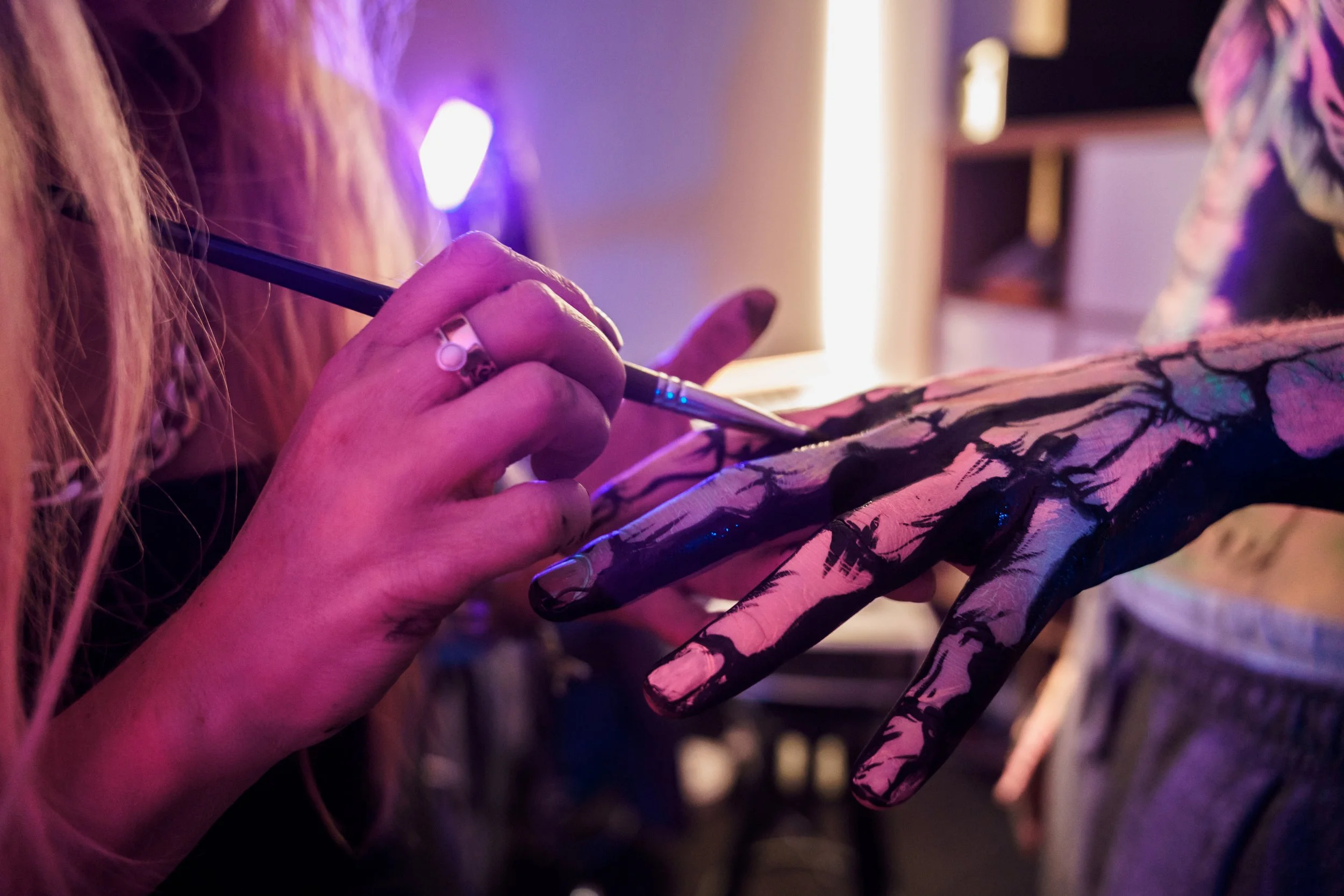 A person is painting black ink or paint on a person's hand with a fine brush, creating a detailed design or tattoo illustration.