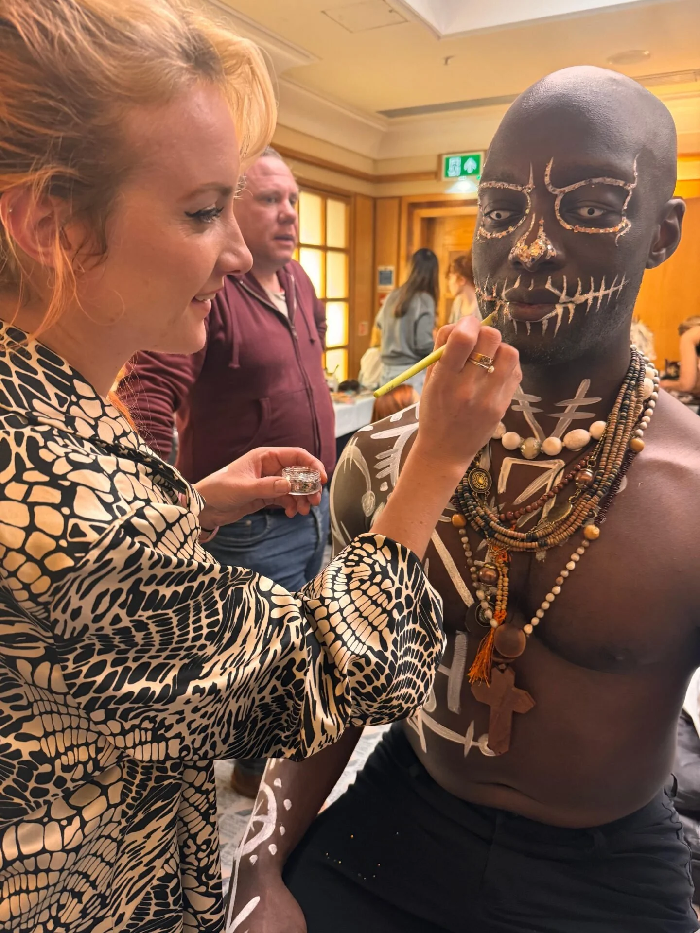 A woman applying face paint to a man with dark skin, who has traditional face paint and necklaces, in a room with wooden walls