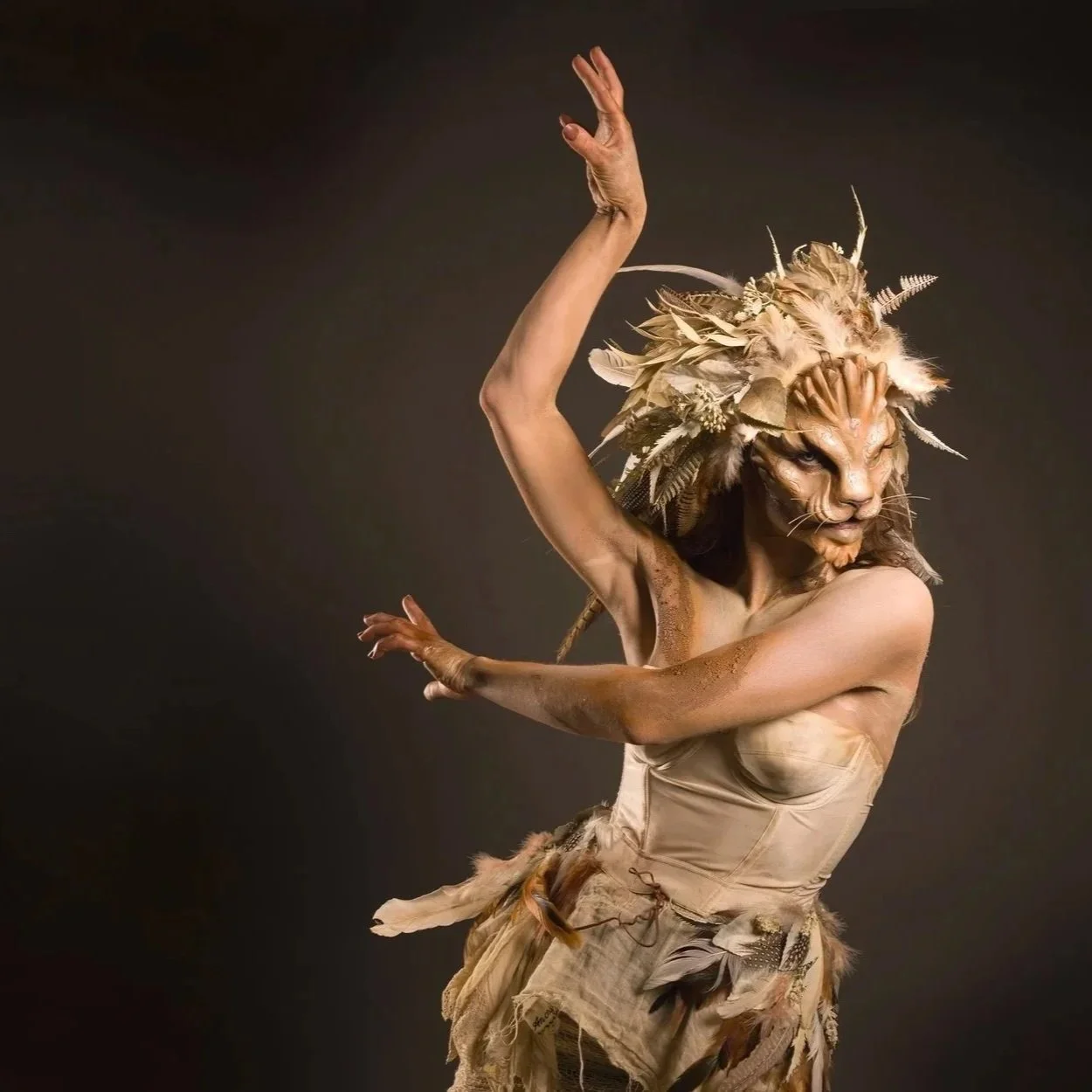 A person dressed in a lion costume with a human body and lion face mask, adorned with feathers and natural elements, posing against a dark background.
