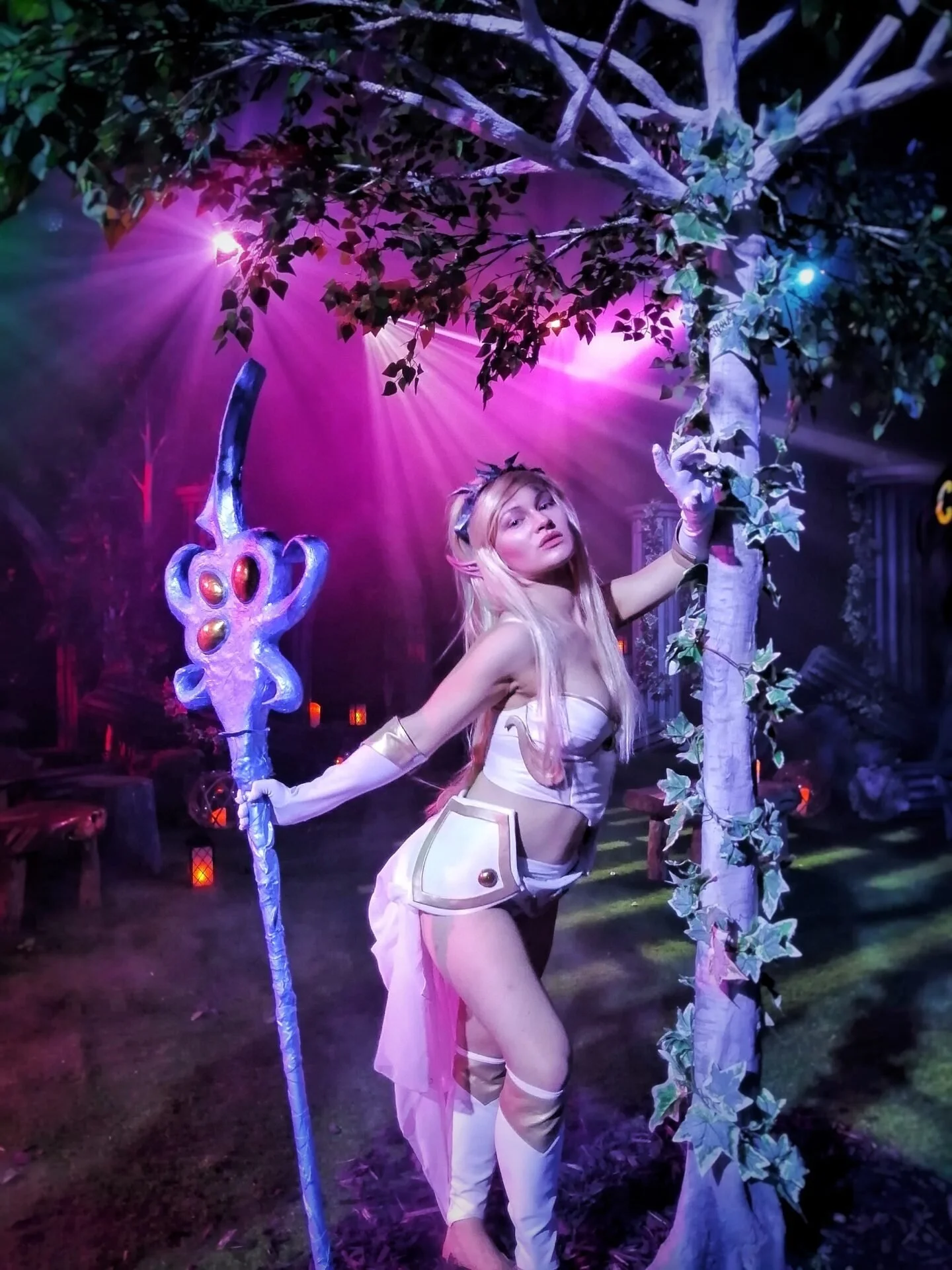 A person dressed in a fantasy costume holding a staff, standing outdoors at night next to a tree with purple and pink lighting in the background.