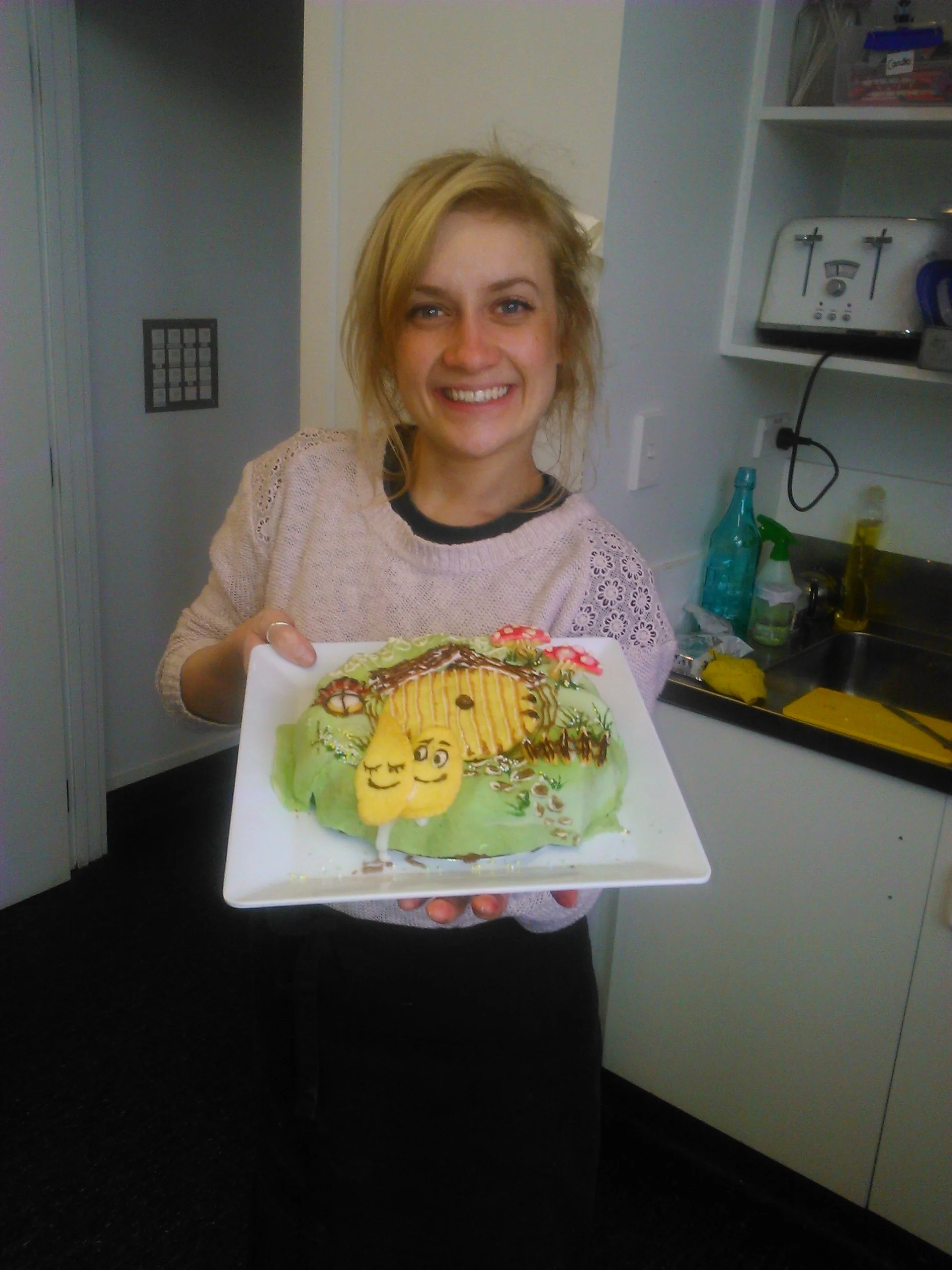 Woman smiling and holding a decorated cake with a house and smiling yellow objects.